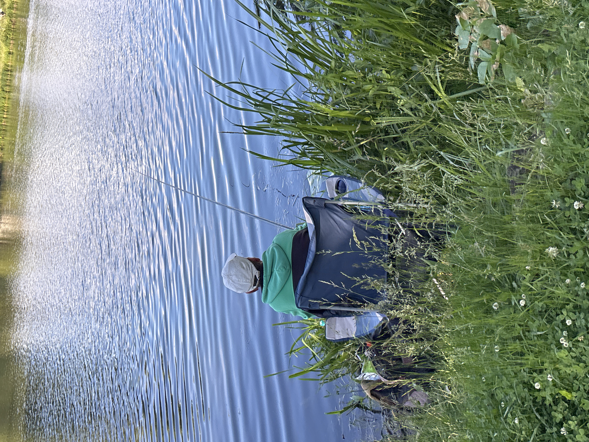 The image depicts a person fishing by a body of water. The person is seated in a chair, facing away from the camera, holding a fishing rod. The environment appears to be a natural setting with lush green grass and reeds surrounding the water's edge. The water surface shows ripples, suggesting a slight breeze. There are no identifiable landmarks to confirm a specific location in Minsk, Belarus.