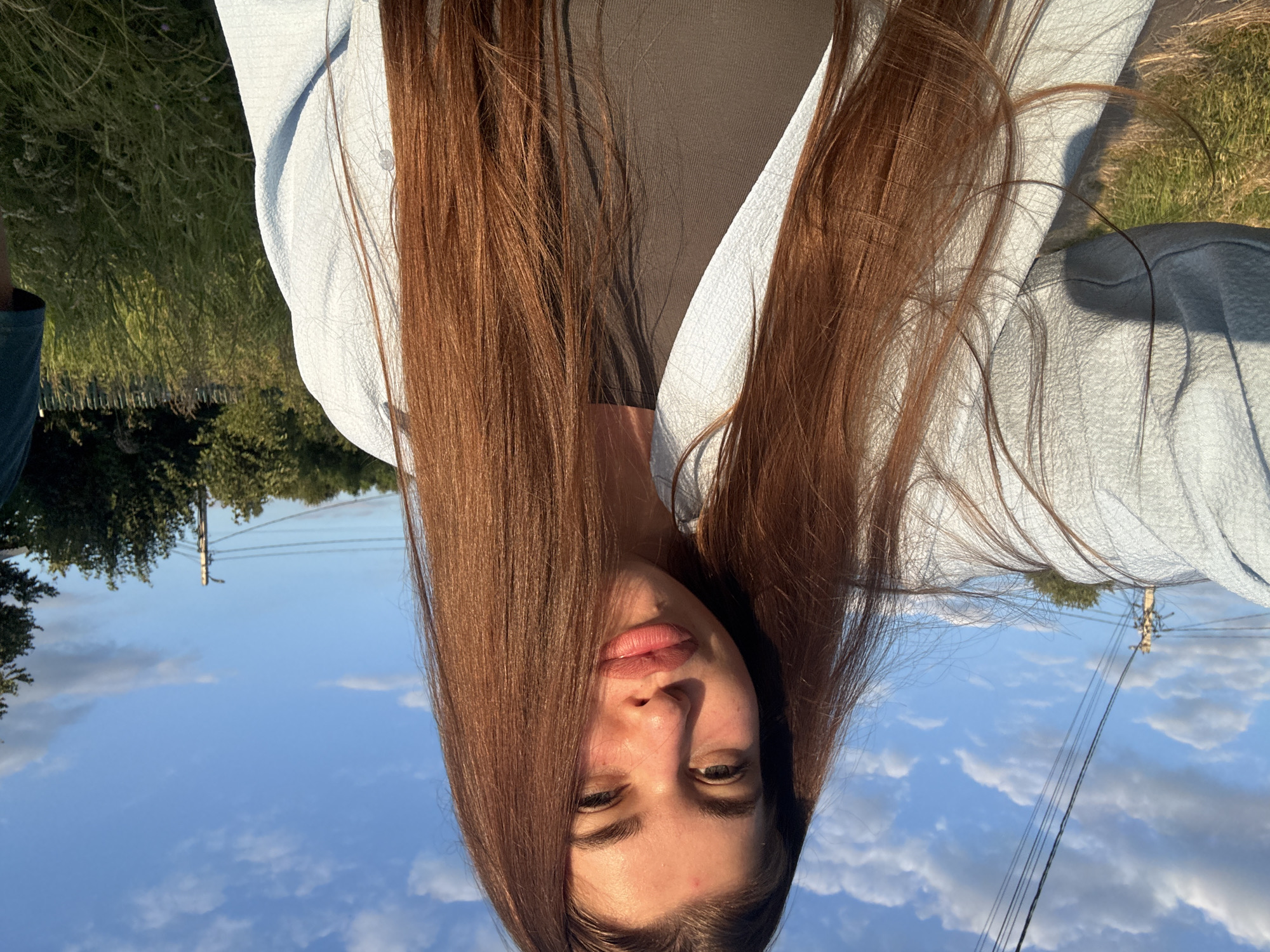 The image shows a young woman with long, reddish-brown hair looking up at the camera. She is wearing a light blue textured top. Her hair is spread out, framing her face and covering part of her clothing. The background is a bright blue sky with scattered white clouds, suggesting it is daytime. There are trees and power lines visible in the distance, indicating an outdoor setting that is likely rural or suburban. The lighting suggests it could be late afternoon due to the warm tones on her skin. The woman's expression is neutral. The photo is taken from a low angle, looking upwards. The scene appears to be set in Мещёры, Russia, as indicated by the location context. There is no visible text in the image.