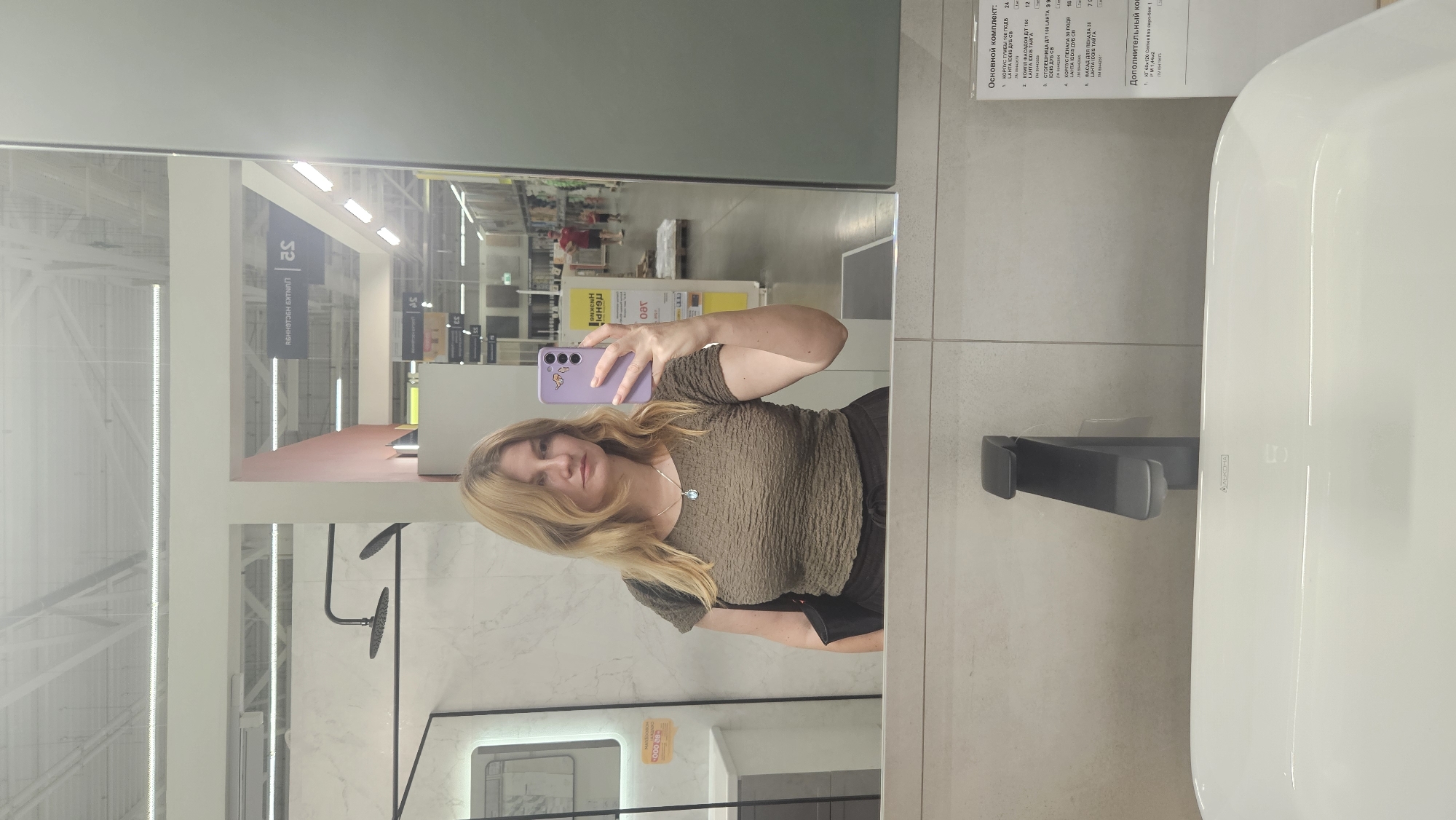 A woman is taking a mirror selfie in a bathroom showroom. She has blonde hair and is wearing a dark olive green ruched top and dark pants. She is holding a purple phone in her right hand. A black handbag is slung over her left shoulder. The background of the mirror reflection shows a well-lit store with aisles of home goods, including shower fixtures and sinks. A price list is visible on the right side of the image, indicating the location is a store in Novaya Adygeya, Russia. A white sink with a black faucet is visible in the foreground.