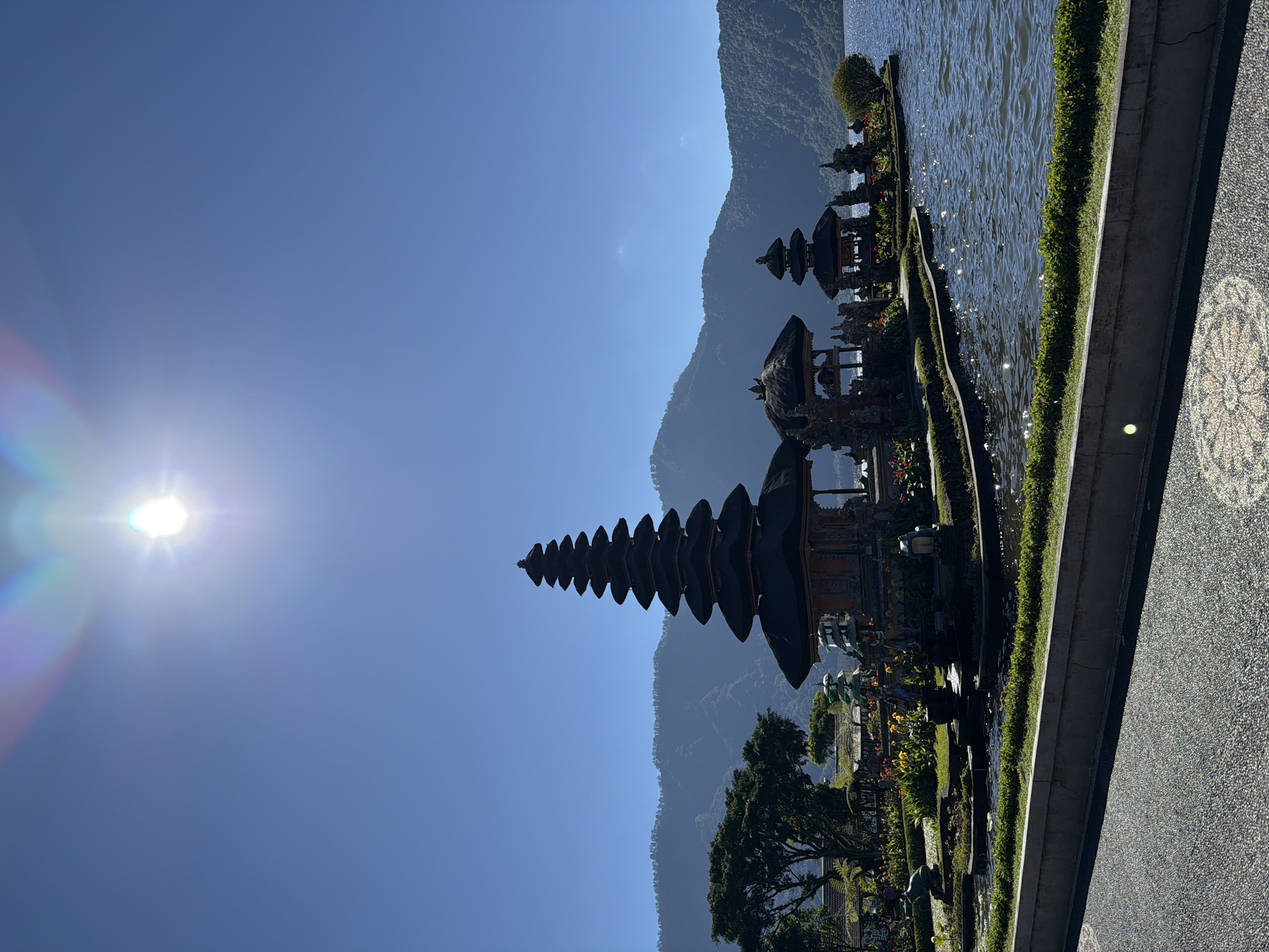 The image captures the Pura Ulun Danu Bratan temple complex situated on the shores of Lake Bratan in the city of Baturiti, Indonesia. The temple, a prominent tourist destination, is characterized by its multi-tiered meru structures, traditional Balinese architecture, and its picturesque setting on a small island within the lake. The bright sunlight, indicated by lens flare in the upper portion of the image, suggests a clear and sunny day. The surrounding environment includes lush green vegetation, trees, and a backdrop of rolling hills covered in dense foliage. The water of the lake is calm with gentle ripples, reflecting the sunlight and the vibrant colors of the temple and its surroundings. In the foreground, a paved walkway borders the lake, with a stone wall and manicured grass separating it from the water. The scene is devoid of any people, focusing entirely on the architectural beauty and natural landscape of the location.