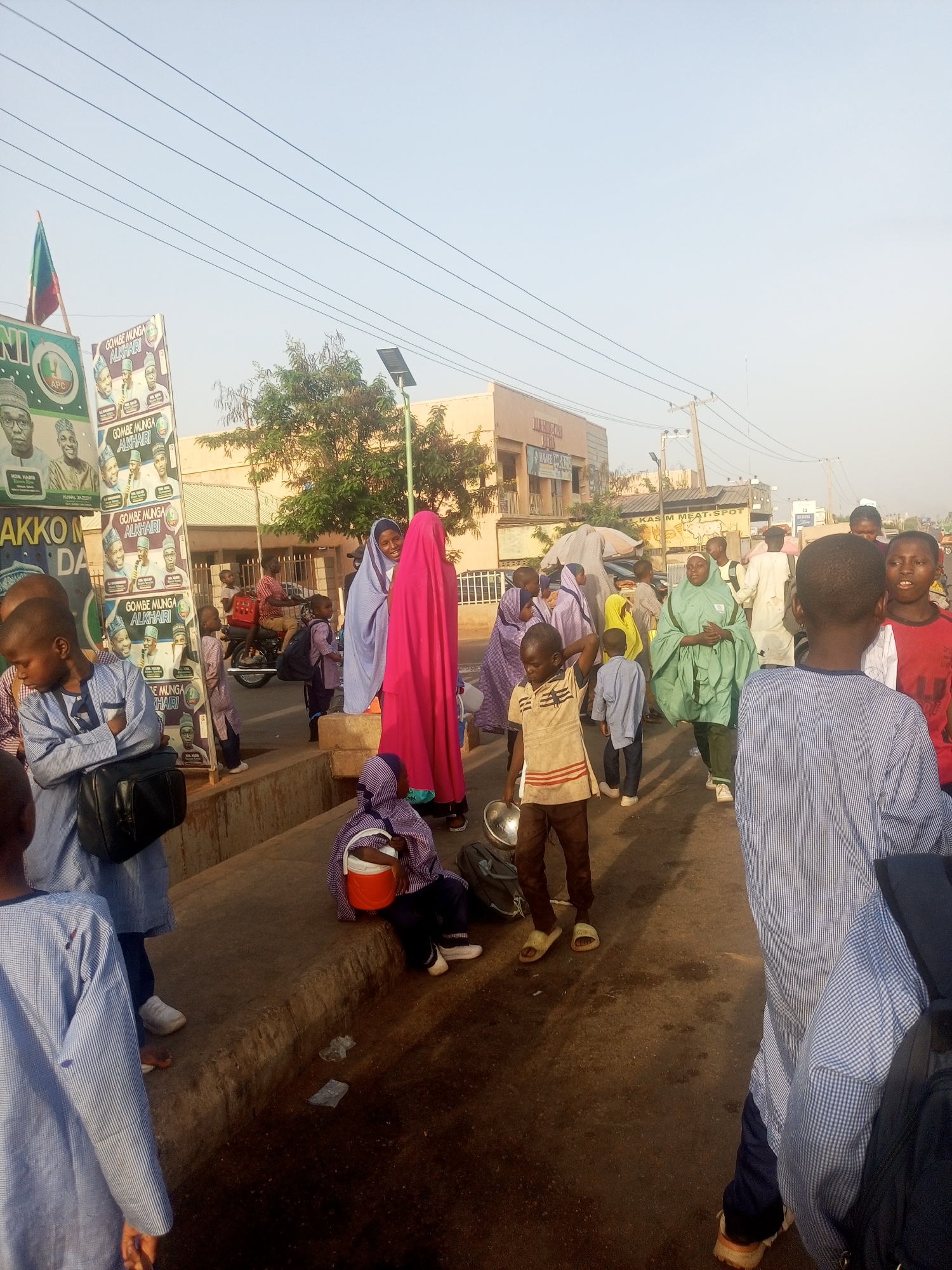 The image shows a bustling street scene, possibly near a school or public gathering place, in what appears to be Unknown, Unknown. A group of children, many wearing similar blue and white striped shirts, are moving along the road. Some carry bags, indicating they may be students.
Several women are visible in the background, dressed in traditional Muslim attire, including hijabs in various colors like purple, pink, and green. A boy in the foreground holds a metal bowl, which may be indicative of some street vending or food-related activity.
In the background, buildings and electrical power lines are visible. There are also several posters on a stand featuring political figures, advertising the "Gombe Munga Alkhairi" campaign. A motorcycle is parked nearby. The scene is bathed in sunlight, suggesting it is daytime.