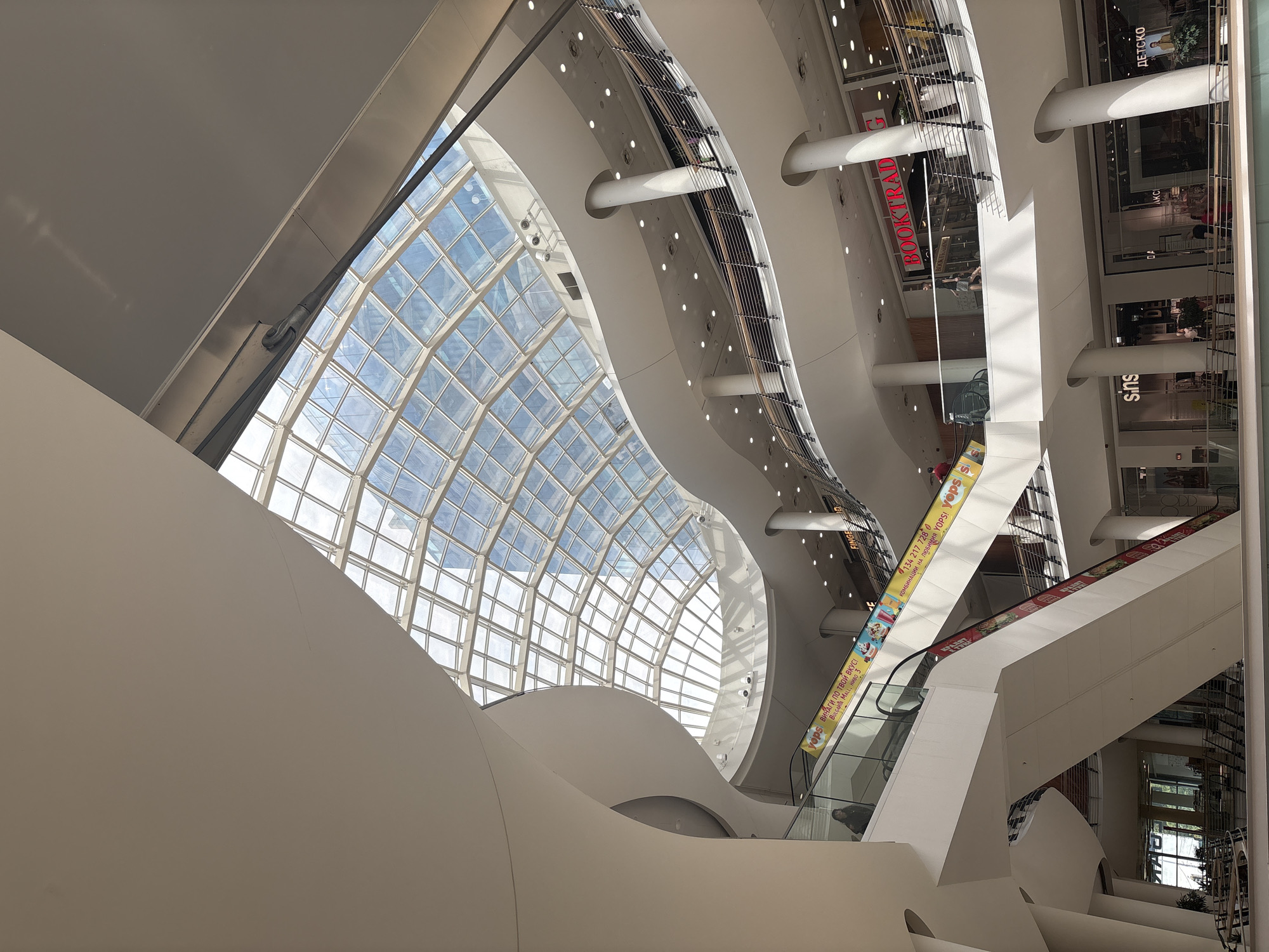 Here is a detailed description of the image:
This is an indoor view of what appears to be a large, modern shopping mall. The architectural design is characterized by flowing curves and large, open spaces. A prominent feature is a curved, grid-patterned glass ceiling, which allows natural light to flood the interior.
The architecture also incorporates white, curved structural elements that contribute to the organic, flowing aesthetic. Various levels and walkways are visible, with escalators connecting different floors. These escalators have advertisements on their sides.
Several store signs are visible, including "BOOKTRAD", "Sins", and a sign in Cyrillic script. There are also advertisements for "YOPS".
The overall environment is clean, modern, and well-lit, suggesting a commercial space designed for shopping and leisure activities.