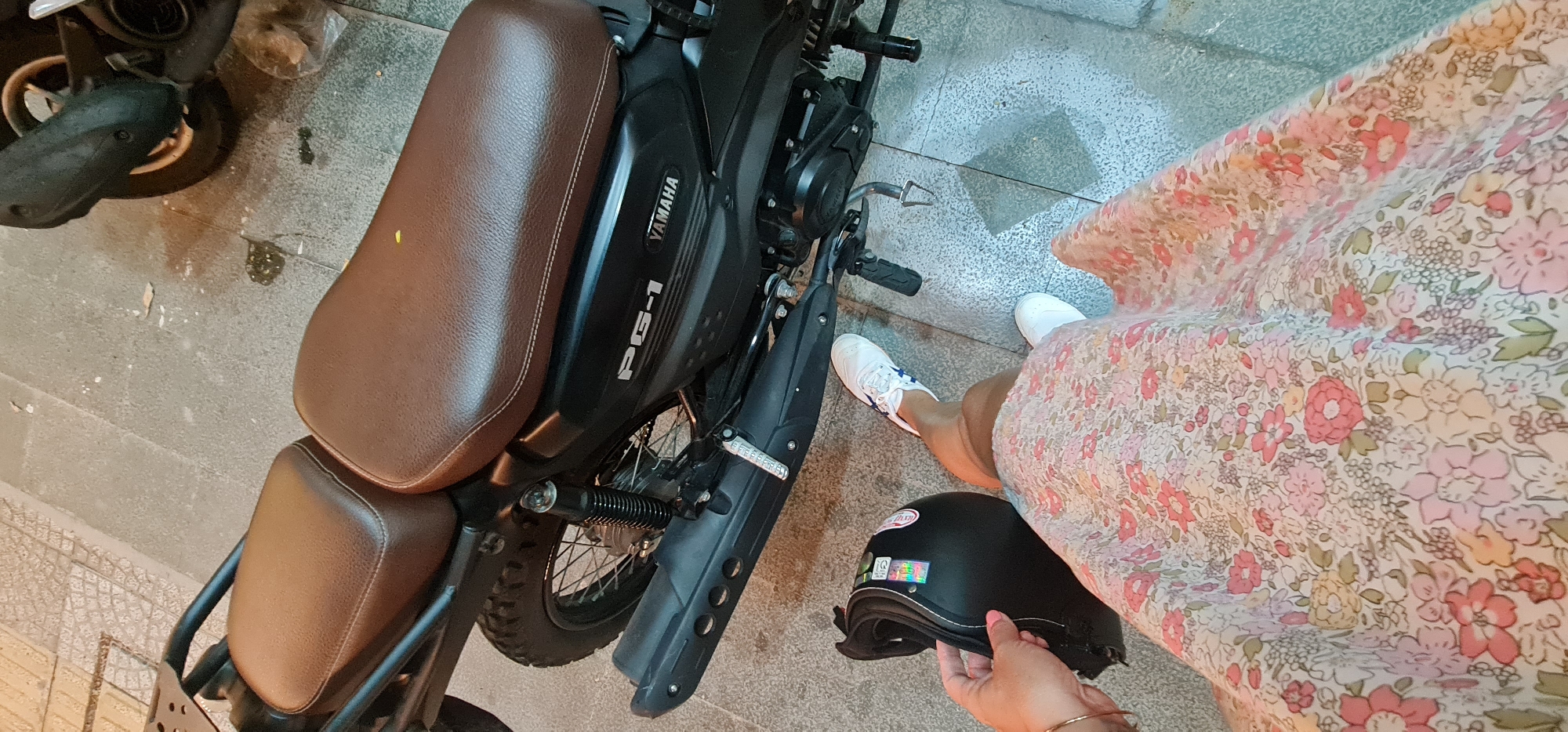 The image shows a black Yamaha PG-1 motorcycle parked on a tiled surface. The motorcycle has a brown leather seat. A person wearing a floral dress and white sneakers stands next to the motorcycle, holding a black helmet in their hand. The environment appears to be outdoors, possibly on a sidewalk or street in a city. Given the presence of the Yamaha PG-1 motorcycle, which is available in Vietnam, and the style of the tiles, the location could be in Đà Nẵng, Vietnam.