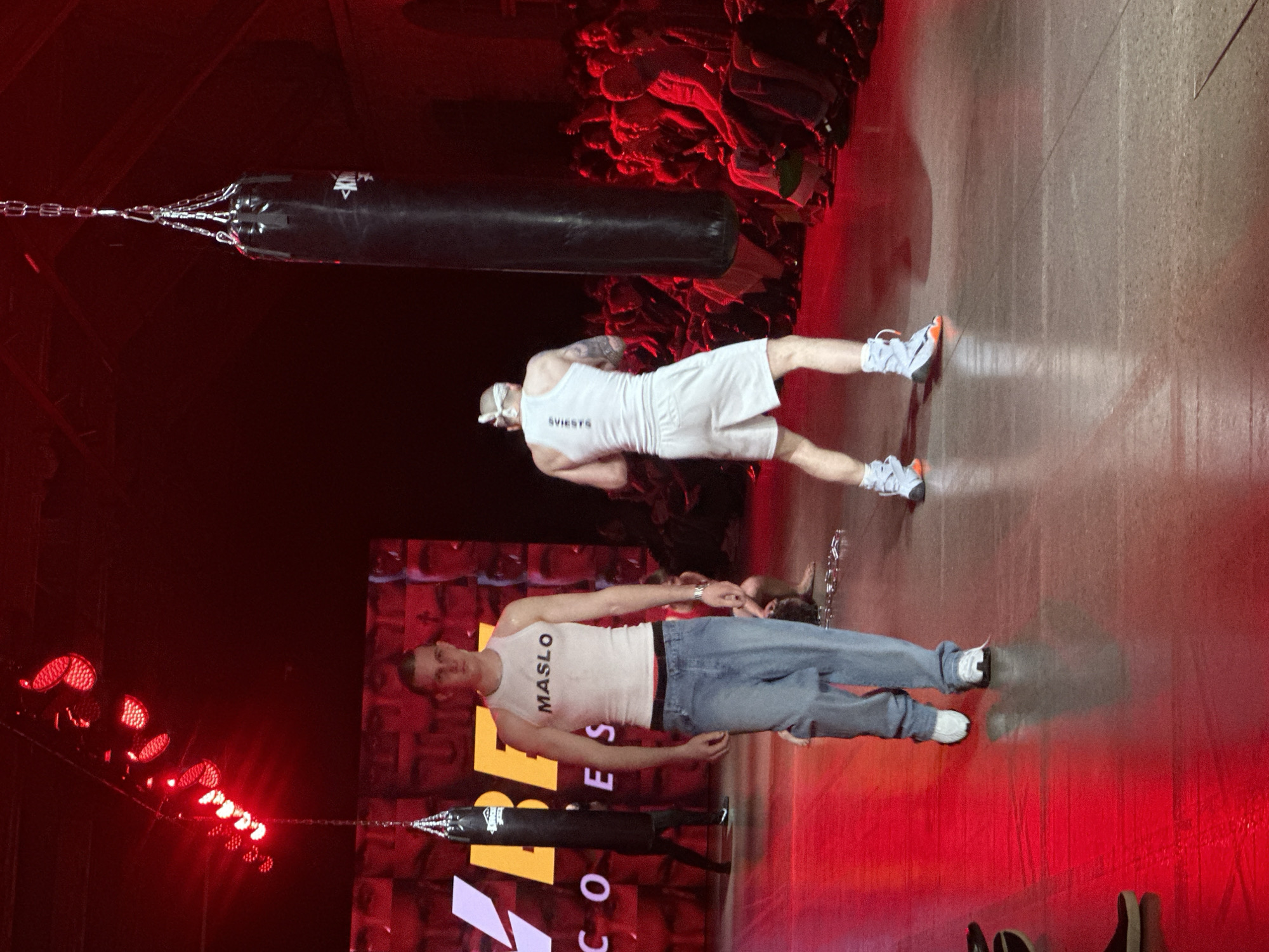 A low-angle, tilted shot reveals a stage setting illuminated by red lighting, possibly a performance or sporting event. Two men are the central figures. One man is wearing jeans, a white tank top with "MASLO" printed on it, and white sneakers. The other man wears a white tank top with "SVIESYS" printed on it, white shorts, a white bandana, and light-colored athletic shoes. Both men are standing on a stage that appears to have a reflective surface. A black punching bag hangs above each man. The image suggests that the men are either preparing to engage with the punching bags or have just finished doing so. In the background, a crowd is visible. A promotional sign can be seen on the back wall. Based on the text visible on the promotional sign "OCO", this image could be taken at the "OCO" venue, a concert venue located in Riga, Latvia.