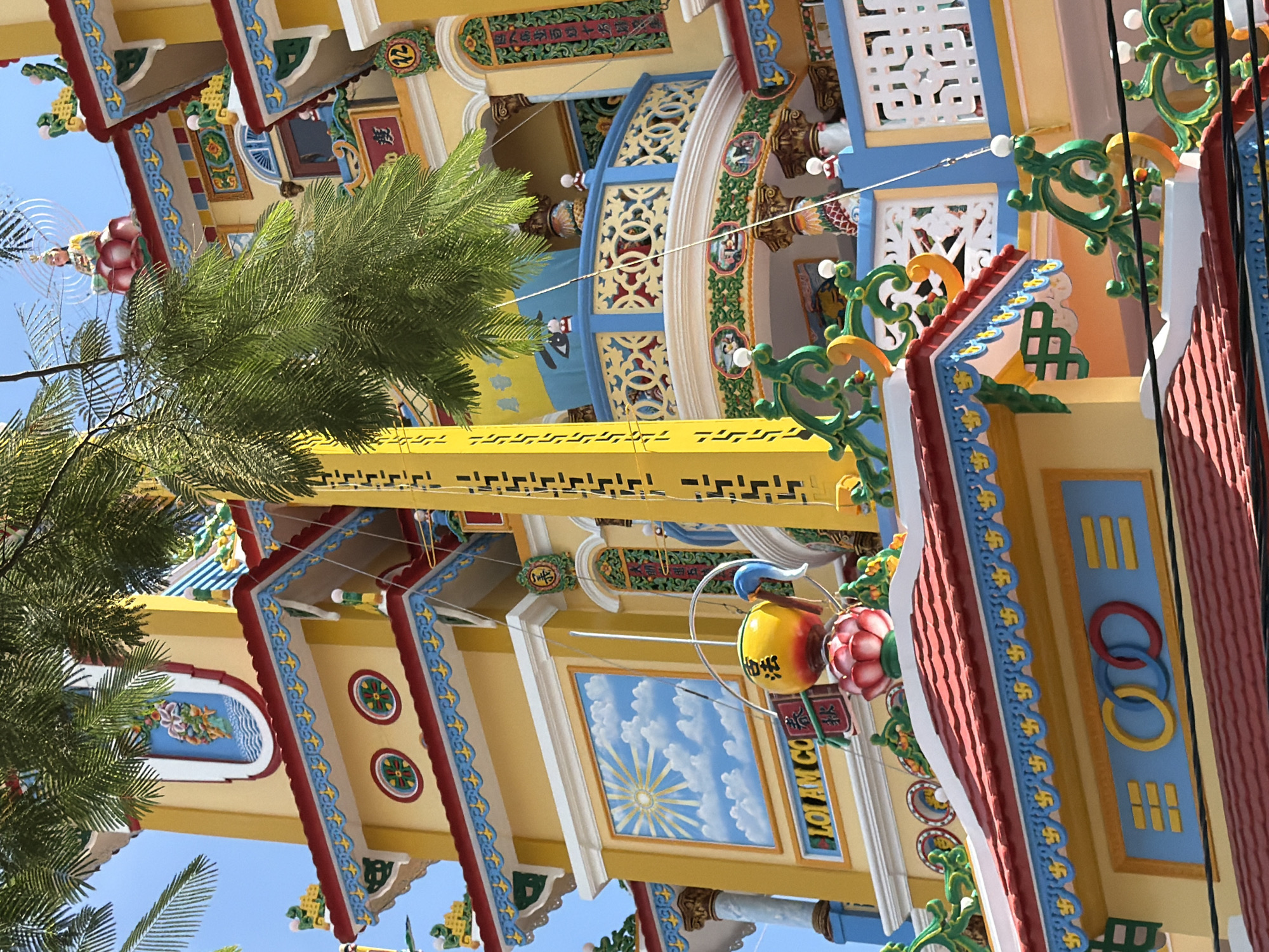 This is a detailed, eye-level shot of a brightly colored temple building with yellow and red accents, possibly located in Nha Trang, Vietnam. The foreground is dominated by a large, green palm frond hanging from above, partially obscuring the view. The temple architecture is intricate and features multiple levels with ornate balconies and decorative elements. On the upper levels, figures of deities or statues are visible, some seated on lotus flowers. The walls are adorned with colorful murals, including one depicting a sun with radiating rays and clouds. There are also decorative carvings and patterns, including what appear to be Buddhist swastikas. A tall, yellow banner with black writing runs vertically along the side of the building. The overall impression is one of vibrant religious and cultural significance.