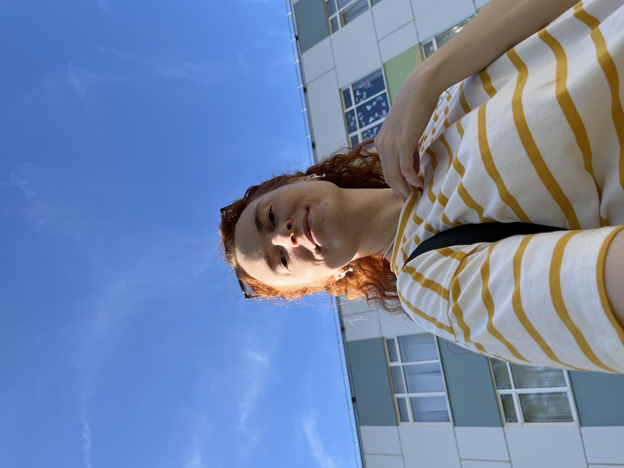 A woman with red curly hair is looking down at the camera. She is wearing a white shirt with yellow stripes and sunglasses perched on her head. Behind her is a clear blue sky with wispy clouds and a building with white and blue paneling and rectangular windows. The location is likely in the city of Chaikovsky, Russia.