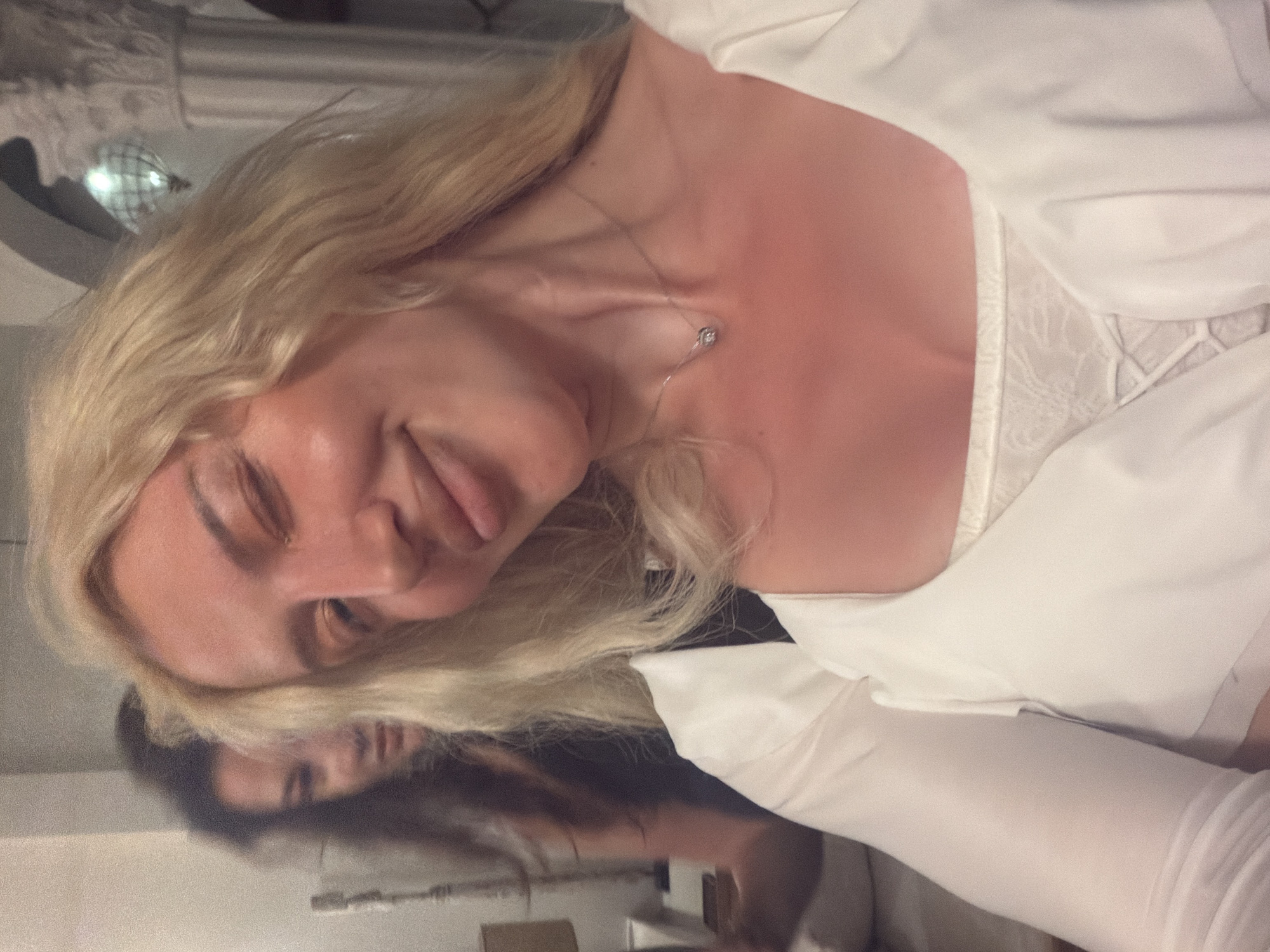 An indoor, vertically oriented close-up photograph captures a person with long blonde hair and fair skin, likely female, wearing a white collared shirt over a white lace undergarment. The subject's head is tilted to the side, with eyes closed or looking downwards, and a playful expression featuring their tongue slightly extended. A delicate silver necklace with a small, round pendant is visible around their neck.
In the background, out of focus, is a softly lit room with what appears to be a decorative, possibly reflective or glass, spherical object hanging. Below the main subject, a reflective surface shows a blurred reflection of a person with darker hair, indicating another individual or the main subject from a different angle. The lighting is dim, suggesting an evening or nighttime setting. No text is visible in the image. The scene conveys a casual and possibly intimate moment, set in North Kuta, Indonesia.
