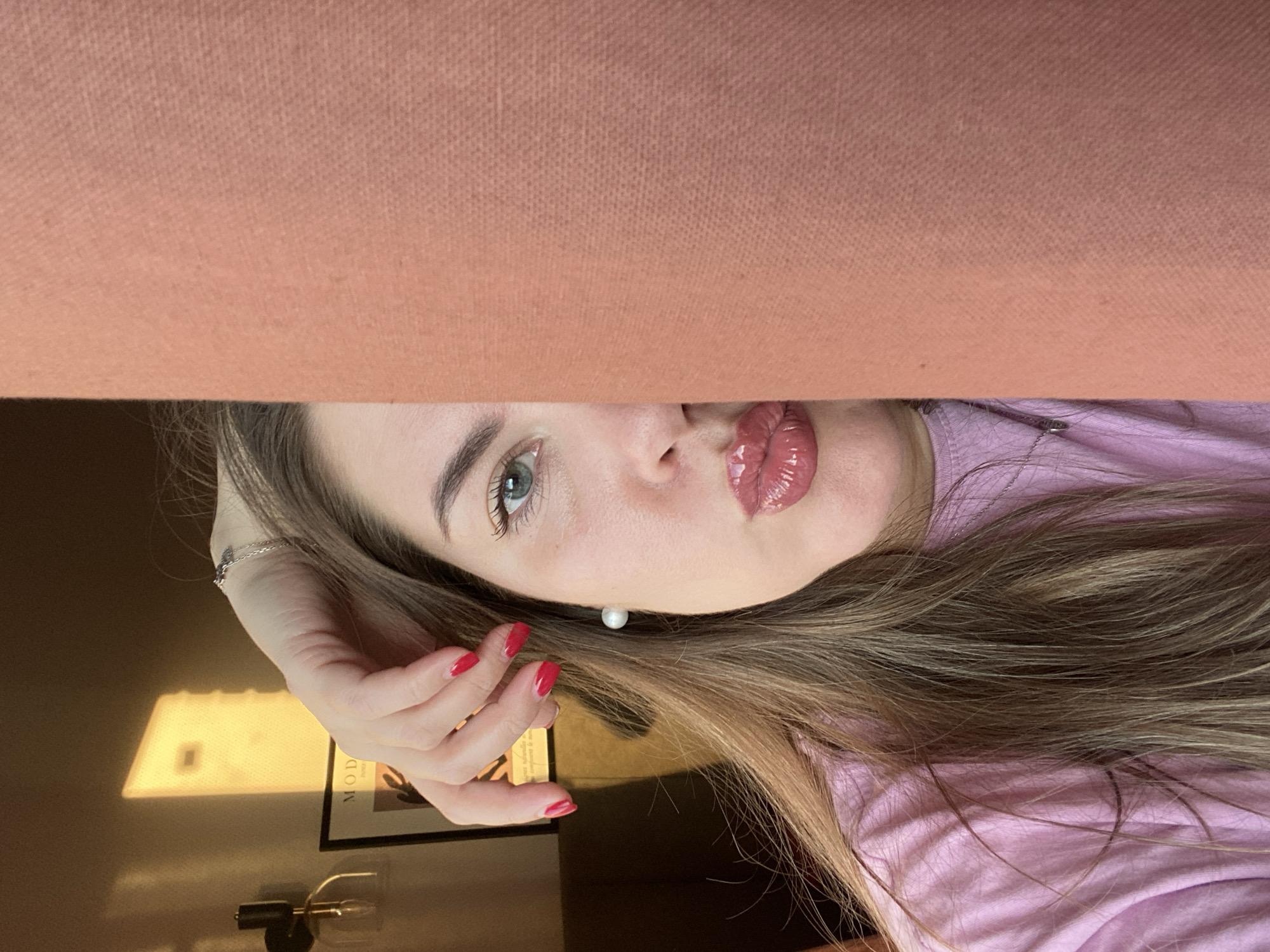The image is an upside-down selfie of a woman. The upper portion of the frame is obscured by a light peach-colored fabric, possibly a blanket or piece of furniture. Only the lower portion of the woman's face, her hair, and one hand are visible. She has light skin, green eyes, and glossy lips. Her hair is long, light brown, and blonde. She is wearing a light pink top. Her fingernails are painted red. In the background, out of focus, there is a lamp and a framed print on a wall. The location is not discernible as being in Izhevsk, Russia, or any other specific city.