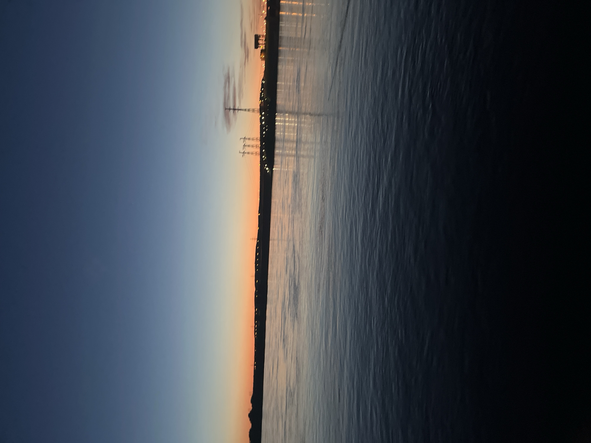 The image depicts a serene twilight or dawn scene over a body of water in Chaikovsky, Russia, captured with a vertical orientation. On the left side, the sky displays a gradient of colors, transitioning from a deep indigo blue at the top left to a softer blue and subtle orange glow towards the center of the image. This color blend suggests the sun has either just set or is about to rise.
A dark, silhouetted landmass or shoreline runs vertically down the middle of the image, separating the sky from the water. Along this distant shoreline, numerous small, bright artificial lights are visible, casting faint, shimmering reflections onto the water's surface. Among the indistinct structures on the shoreline, a prominent tall transmission tower or power line pylon with visible crossbars stands out.
The right side of the image is dominated by the dark, rippled surface of the water, likely the Kama River or a associated reservoir, reflecting the dim light from the sky and the artificial lights on the shore. No human activity or specific events are depicted, and no text is visible. The calm waters and clear sky contribute to a peaceful and atmospheric setting.