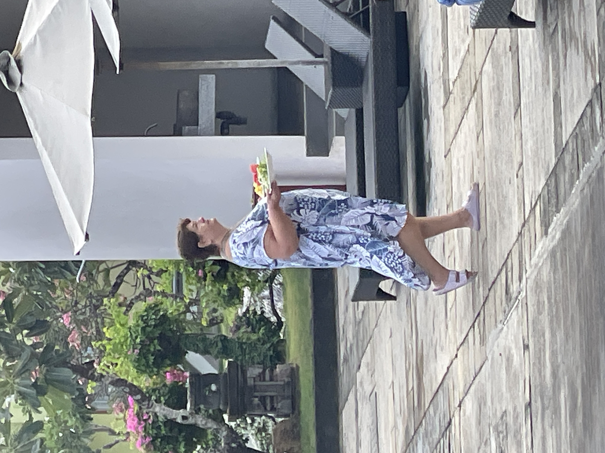 A woman in a blue and white floral dress walks on a paved outdoor area, carrying a white plate with fruit salad. She is walking towards the right side of the frame. Behind her, there is lush greenery with pink flowers, a stone sculpture, lounge chairs, and an umbrella. The scene appears to be at a resort or hotel in Kuta Selatan, Indonesia, likely near a pool area given the paving stones and lounge chairs.