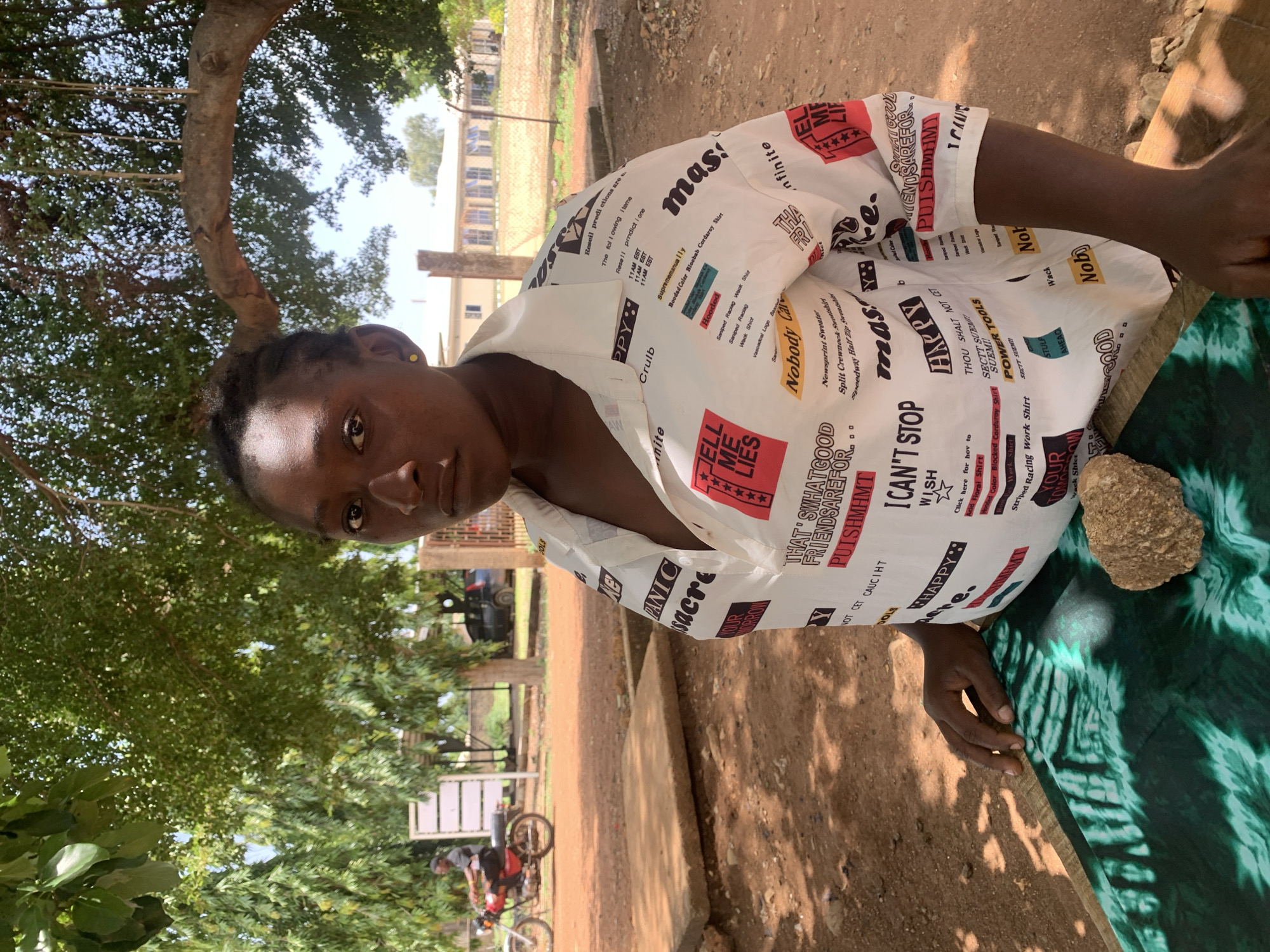 A person with dark skin and short dark hair, wearing a white shirt with printed words, is seen outdoors under a tree. The shirt has phrases like "TELL ME LIES", "I CAN'T STOP", "Nobody Car", and "POWER TOOLS". The person is looking toward the camera. A brown rock is visible on a green and black patterned surface. In the background, there is a dirt road, a building with a fence, and a person on a bicycle. The image was taken in Maigizo, Nigeria.