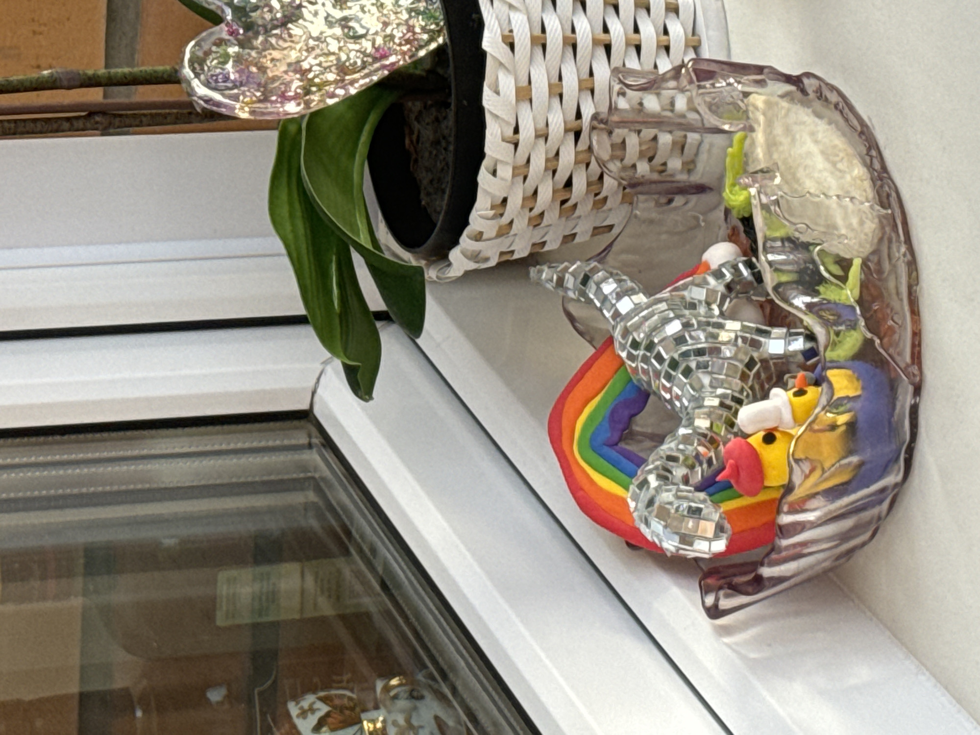 A close-up shot depicts a windowsill with various objects. A houseplant in a white woven basket with a black inner pot sits on the left side. To the right of the plant is a decorative object consisting of a mirror-covered reptile figure reclining on a rainbow. The rainbow and reptile figure are encased within a clear plastic or glass shape. The windowsill itself is white and sits above a window with a visible frame. The location is Krasnoyarsk, Russia.
