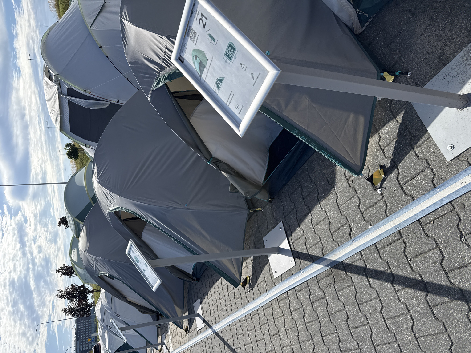 This image depicts an outdoor display of tents in the city of Bielany Wrocławskie, Poland. The tents, primarily gray and light blue, are set up on a paved surface, likely a parking lot or an open plaza. Several display stands are placed in front of the tents, featuring information and diagrams about the products. In the background, a multi-story building and trees suggest an urban or suburban environment. The sky is partly cloudy, indicating daytime. There are no people visible in the scene, but a small bird is present near the base of one of the display stands. The tents appear to be new and arranged in a neat, orderly fashion, suggesting a commercial exhibition or retail display. The ground is paved with interlocking concrete blocks, and a metal strip runs along the edge of the display area.
