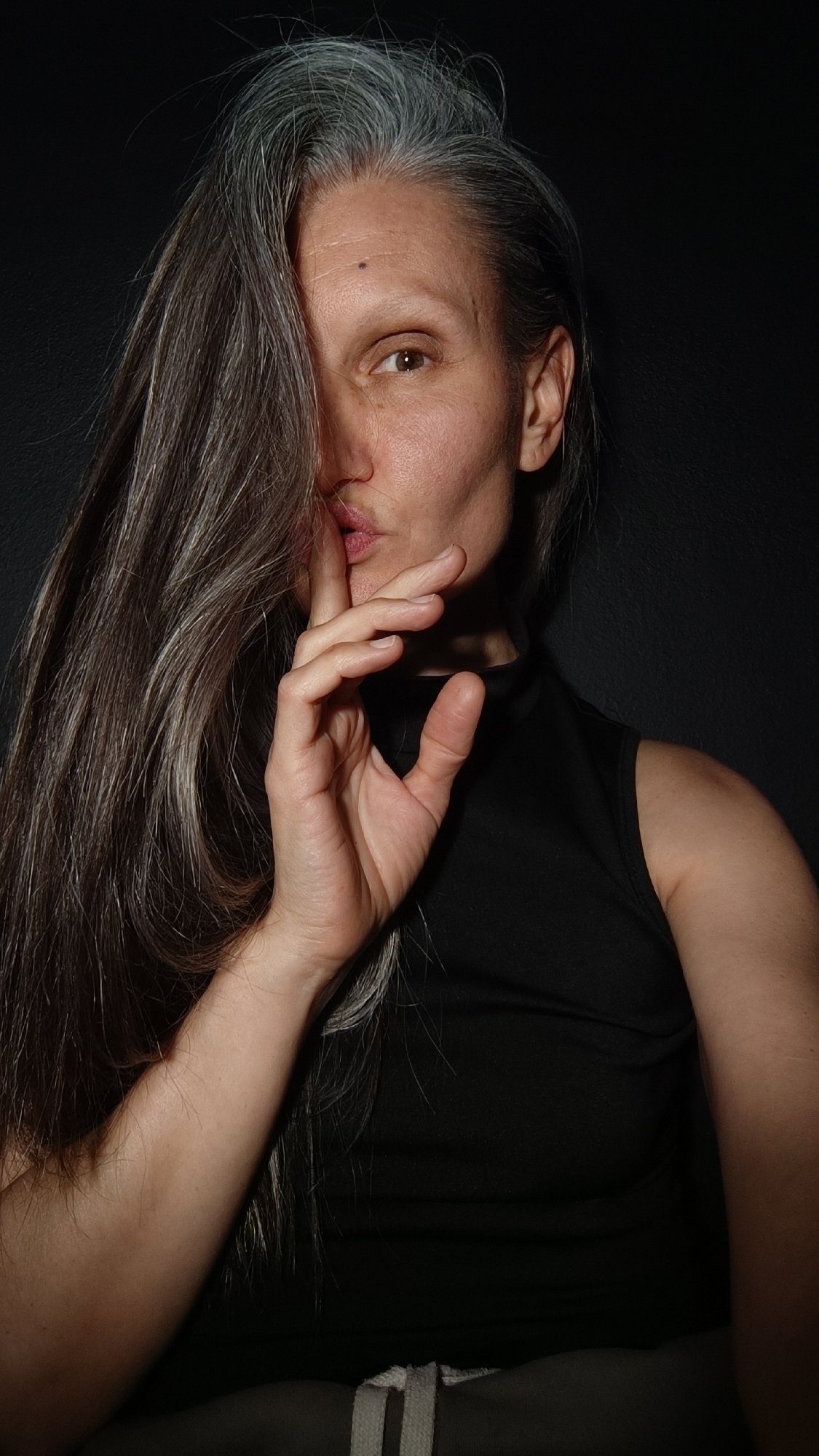 A woman with long, graying hair is positioned against a dark background. Part of her face is obscured by her hair. She is making a shushing gesture with her hand near her mouth. She wears a black sleeveless top. The bottom of the image shows what appears to be a light-colored drawstring. There is no indication in the image to determine if the location is in the city of Lermontov, Russia.