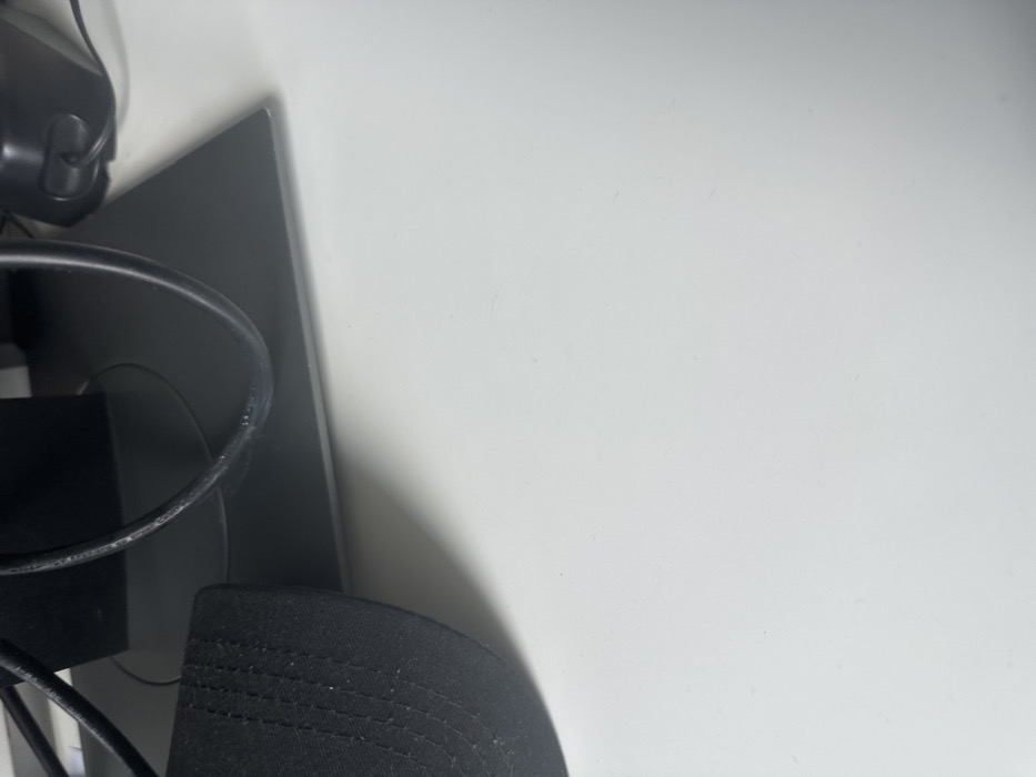 A close-up, top-down view of a white desk surface, located in Thiais, France. In the upper left quadrant, the brim and a portion of the crown of a black fabric baseball cap are visible, exhibiting visible stitching. Centered along the top edge, the dark grey rectangular base of an electronic monitor stand is present. Multiple black electrical cables are routed across and behind this stand; one cable displays white scuff marks. To the upper right, a section of a black electronic device, consistent with a computer speaker featuring a dark grill and a circular button, is partially observed. The desk surface is uniformly lit, casting faint shadows from the objects. No individuals are present, and no actions or interactions are depicted.