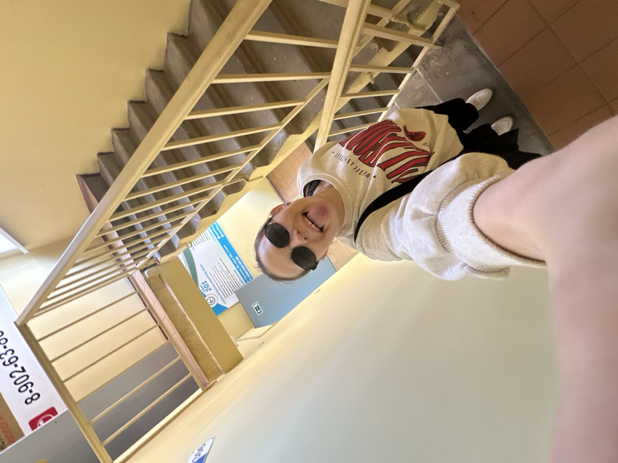 The photograph appears to be a selfie taken by a young woman standing on a tiled landing or hallway in a building with a staircase and cream-colored walls. She is wearing black pants, a light-colored hooded sweatshirt with a design, and white shoes. She also has on black sunglasses and is sticking her tongue out in a playful manner.
The staircase has a light wood or metal railing. A sign on the wall behind her advertises office 201, referencing "ПЛАСТИЧЕСКАЯ ХИРУРГИЯ ЛИЦА" (plastic surgery). The phone number 7 (342) 219-09-89 and an address, ul. Pushkina, 30, office 201, 2 etazh, are visible, indicating a location in Perm, Russia. There is also a phone number "8-902-63..." on a sign to the left.