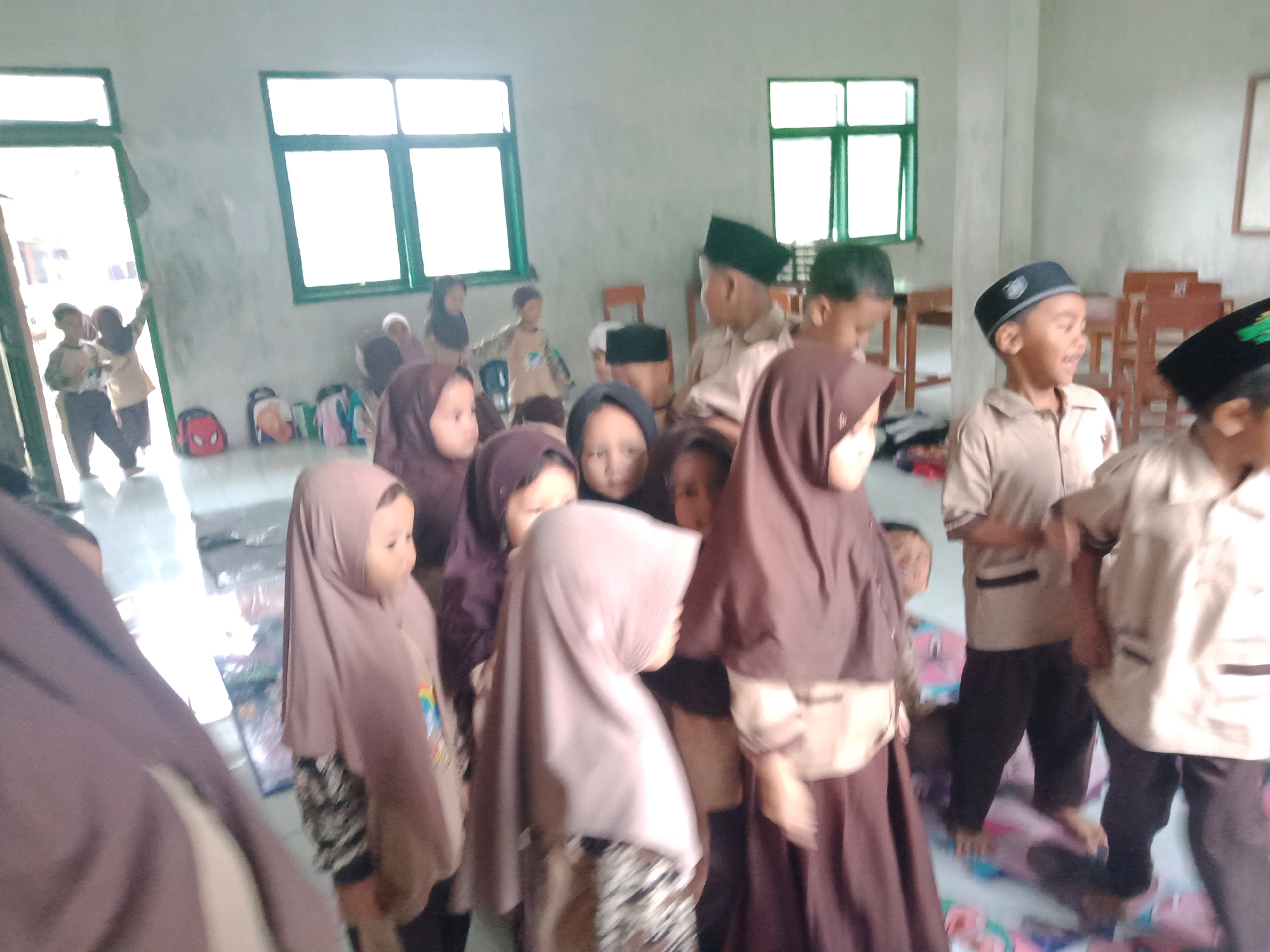 In Cilampunghilir, Indonesia, a group of young children, both boys and girls, are gathered indoors. Many of the children are wearing hijabs or peci caps, suggesting a religious setting, possibly a school or prayer area. Some children are wearing uniforms, consisting of light-colored shirts and dark pants. Backpacks, including one with a Spider-Man design, are visible on the floor near an open doorway. The room has windows with green frames and light-colored walls. The scene appears to be a candid moment, possibly during a class, a break, or a prayer activity.