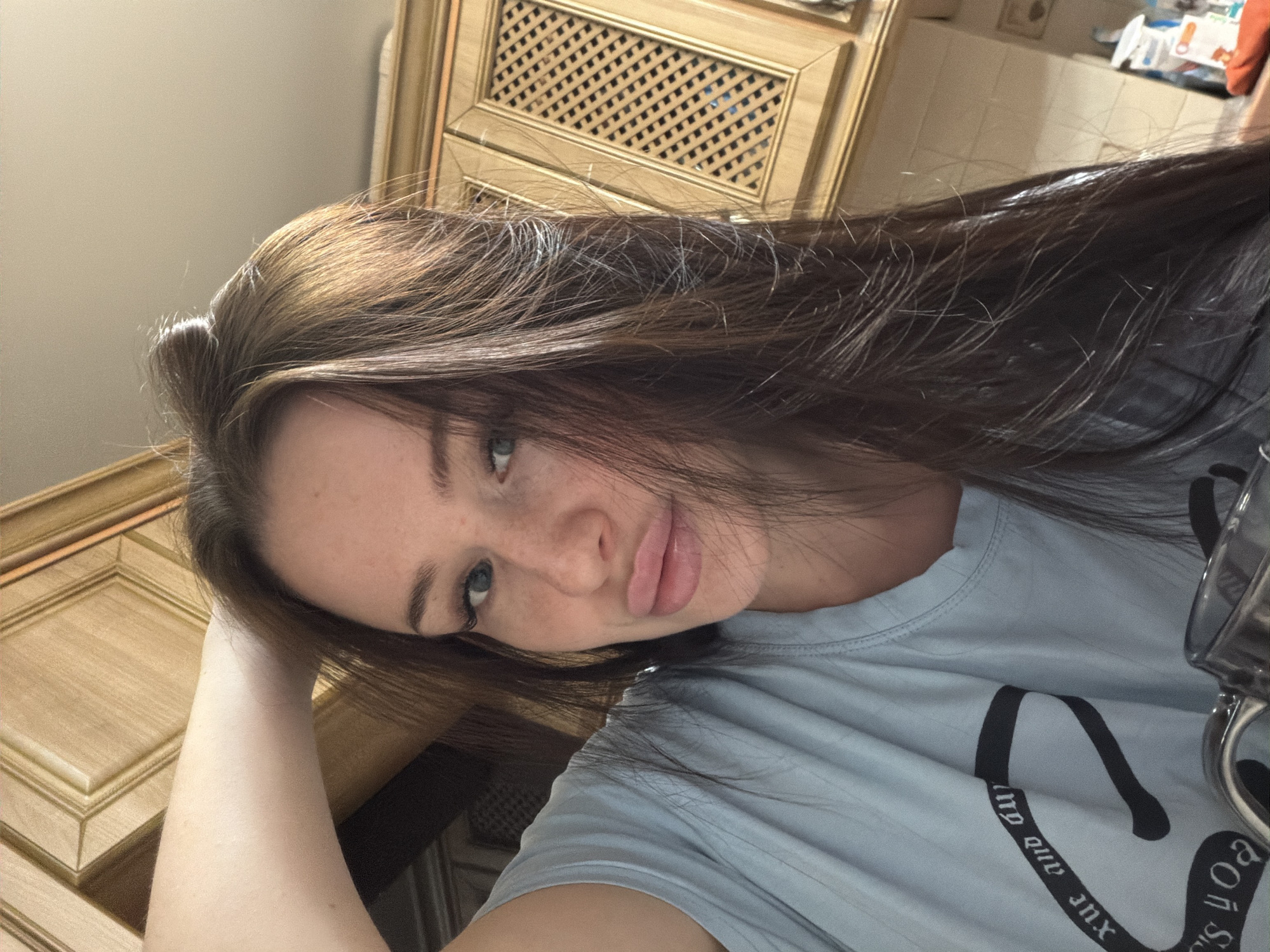 A young woman is depicted in a close-up, casual, selfie-style shot, lying down or reclining with her head tilted to the left. She looks towards the camera with a playful, slightly pouty expression, her lips slightly parted. Her left arm is visible, bent at the elbow, appearing to support her head. She has long, dark brown hair with lighter, possibly highlighted, streaks, and blue-green eyes. Freckles are visible across her nose and cheeks. She wears a light blue t-shirt.
The setting appears to be indoors, likely a home, with wooden kitchen or dining cabinets forming the background. One cabinet features a decorative latticework panel. The background is somewhat out of focus, suggesting the photo was taken with a shallow depth of field. A silver-colored object, possibly a mug or part of a pair of glasses, is partially visible on the right edge of the frame.
The light blue t-shirt has black text printed on it, partially visible in several places. Fragments include "xp" or "xuc", "and Qu", and "e of yo", suggesting a phrase like "xoxo and Queen" or a similar design. The overall mood is relaxed and candid. The location context is unknown, Russia.
