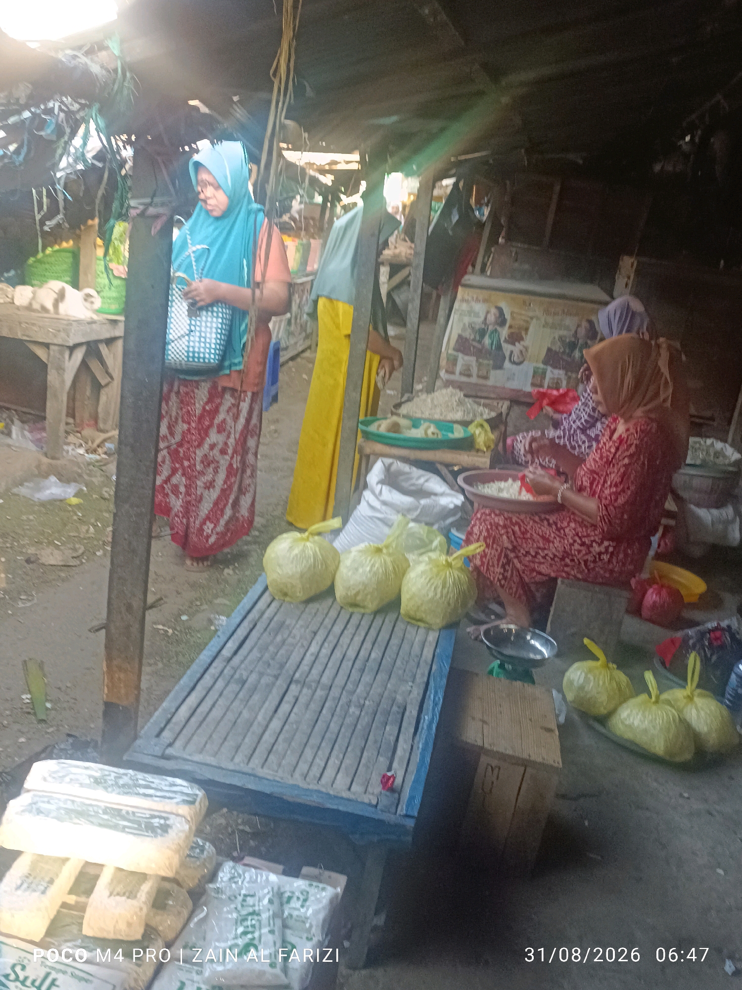 In Arosbaya, Indonesia, a market scene unfolds beneath a makeshift roof constructed from corrugated metal and wood. A woman wearing a turquoise hijab and a red and white patterned skirt stands next to a wooden table, holding a woven bag. Beside her, a person in a yellow garment is partially visible. Further into the market, another woman, draped in a brown hijab and a red patterned dress, sits on a low stool, sorting through what appears to be white food ingredients in a large round bowl. Two other individuals, also wearing hijabs, are visible behind her, one in a purple and white patterned top. In the foreground, a wooden platform covered with bamboo matting displays several large, clear plastic bags filled with yellow produce, possibly fruits like bananas or mangoes. To the left, stacks of packaged goods, some wrapped in plastic with visible text, are arranged. A small silver weighing scale sits on a wooden crate. The ground is dirt and scattered debris. The timestamp "31/08/2026 06:47" is overlaid on the image, along with the text "POCO M4 PRO | ZAIN AL FARIZI".