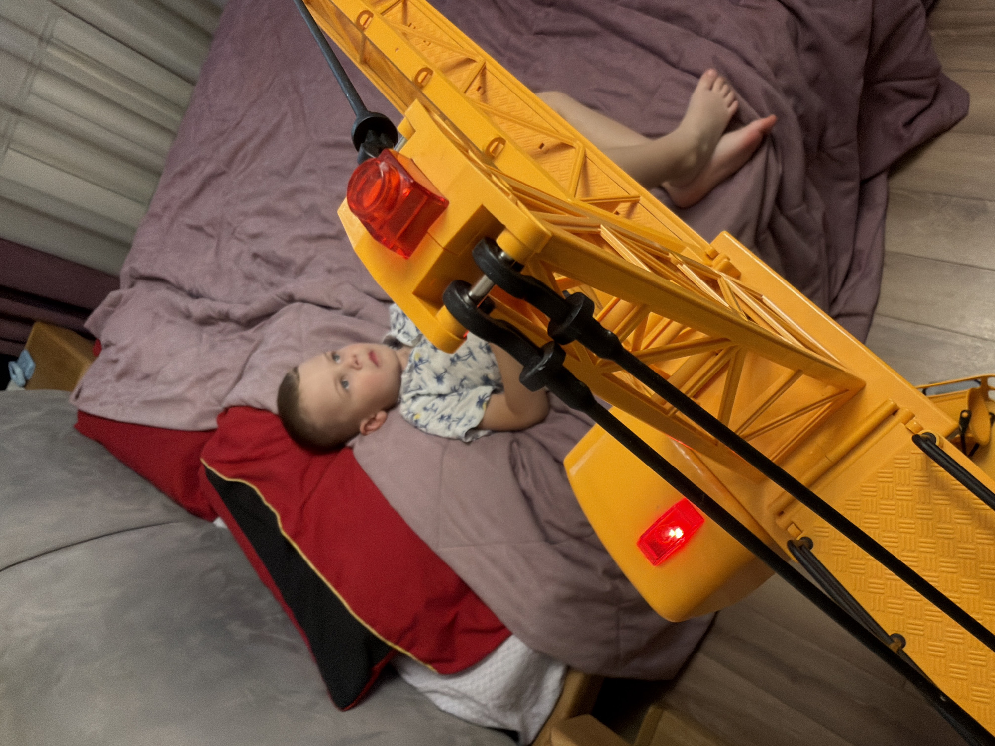 A young boy lies on a purple blanket on a bed. He is wearing a white shirt with a blue pattern. A large yellow toy crane with a red light is positioned in the foreground, partially obscuring the boy. The background shows a grey sofa with red and black pillows. The scene appears to be indoors, likely in a bedroom or living area. The image does not provide any specific visual cues to determine the location within the city of Minsk, Belarus.