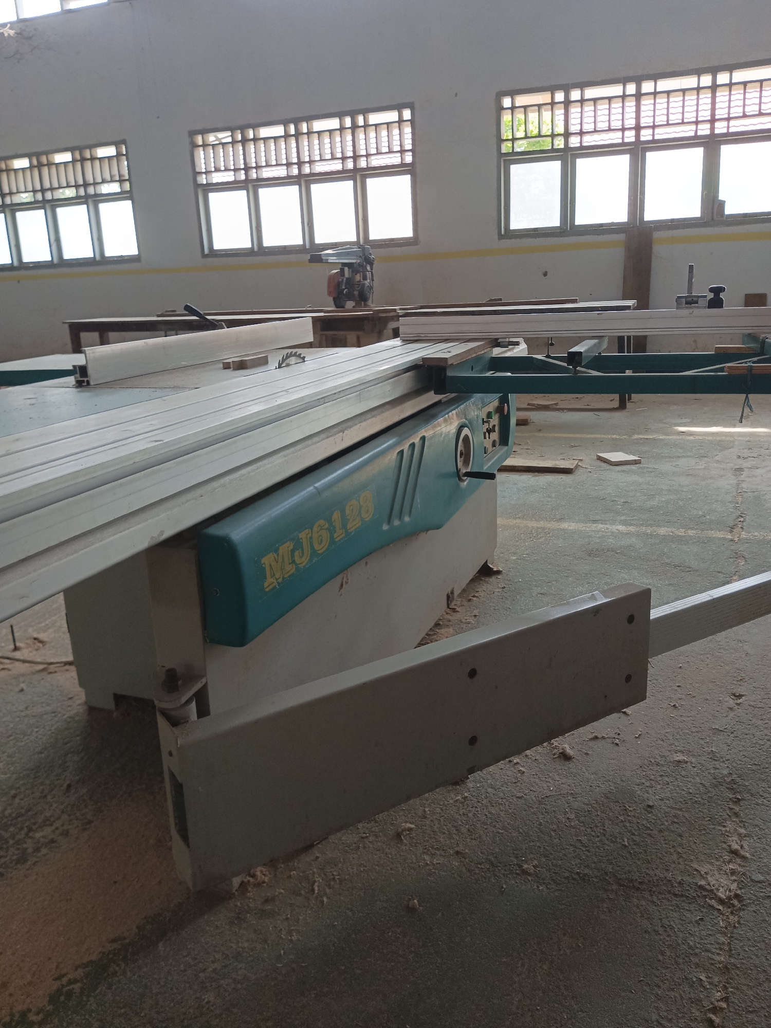 The image depicts a woodworking shop in Dupi, Indonesia. The focal point is a large sliding table saw, identified by the model number "MJ6128" printed on its teal-colored housing. The saw is positioned on a concrete floor covered with sawdust, indicative of ongoing work. In the background, several large windows with louvered upper sections and clear lower panes allow natural light to enter the space. The walls are painted white, and a yellow stripe runs horizontally across them. Various pieces of lumber and other woodworking equipment, including a smaller, portable saw, are visible in the scene. The overall impression is of a functional, albeit somewhat untidy, workshop environment.