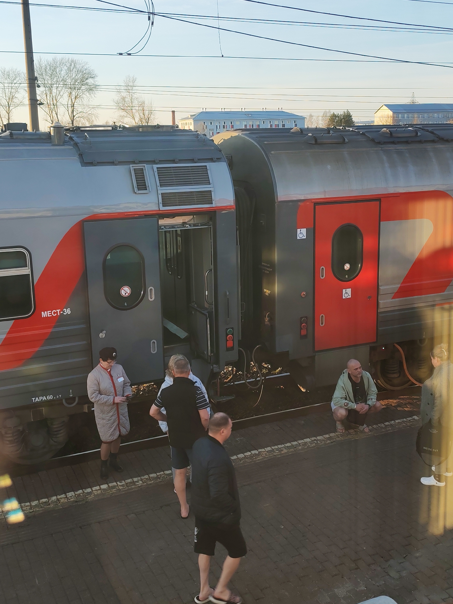 The image captures a train stopped at a platform in Agryz, Russia, bathed in the low-angle light of either early morning or late afternoon. The main subject is a grey and red train car, part of the Russian Railways (RZD) fleet, with one door open, revealing the interior, and another red door closed.
Several people are on the brick-paved platform. A woman in a light-colored coat, possibly a train attendant, stands near the open door, seemingly checking something in her hands. Next to her, a man in a black t-shirt and shorts stands. Further right, a bald man in a light green jacket and white shorts is squatting next to the train. Another person in a grey jacket stands nearby, partially obscured. In the foreground, a man in a black jacket and shorts walks away from the camera.
Visible text on the train includes "MECT-36" (Мест-36, likely indicating "Seats-36") and "ТАРА 60, 3Т" (Tare 60, 3T). Both doors feature no-smoking symbols, and the closed red door displays two wheelchair accessibility symbols. A partial view of a bicycle frame is seen on the ground near the open door. Overhead power lines crisscross the clear sky. In the background, bare trees and various buildings, suggesting a semi-industrial or residential area, are visible. One person carries a bag with "INSIDE OUT" printed on it. The scene conveys a calm, routine stop at a train station.