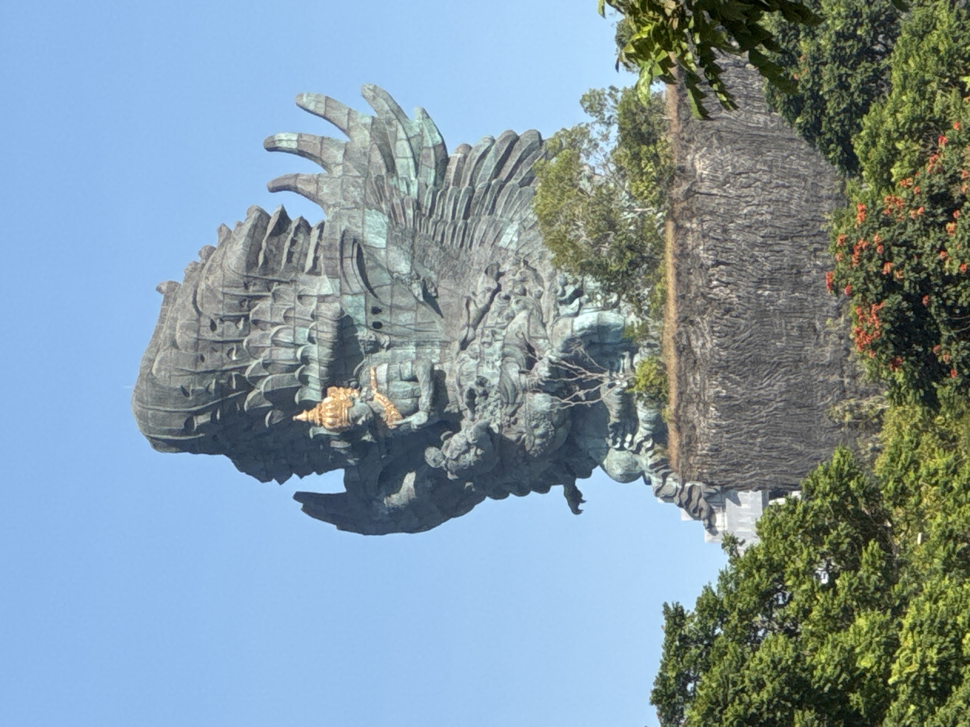 The image displays the monumental Garuda Wisnu Kencana statue, situated in Ungasan, Kuta Selatan, Bali, Indonesia. The central subject is a colossal statue, predominantly bronze-green with a weathered patina, depicting the Hindu deity Vishnu, recognizable by a prominent golden crown and golden chest adornments, mounted on the mythical eagle Garuda. The statue's large, textured wings spread widely, occupying significant portions of the mid-ground and upper-right. It is elevated on a substantial, dark grey, vertically striated cliff-like formation or constructed base. The lower portion of the image features dense green foliage, comprising various trees and bushes, some exhibiting small clusters of reddish-orange blossoms. The upper background is a clear, unblemished blue sky. No human figures or other significant structures are discernible beyond the statue and its immediate environment.