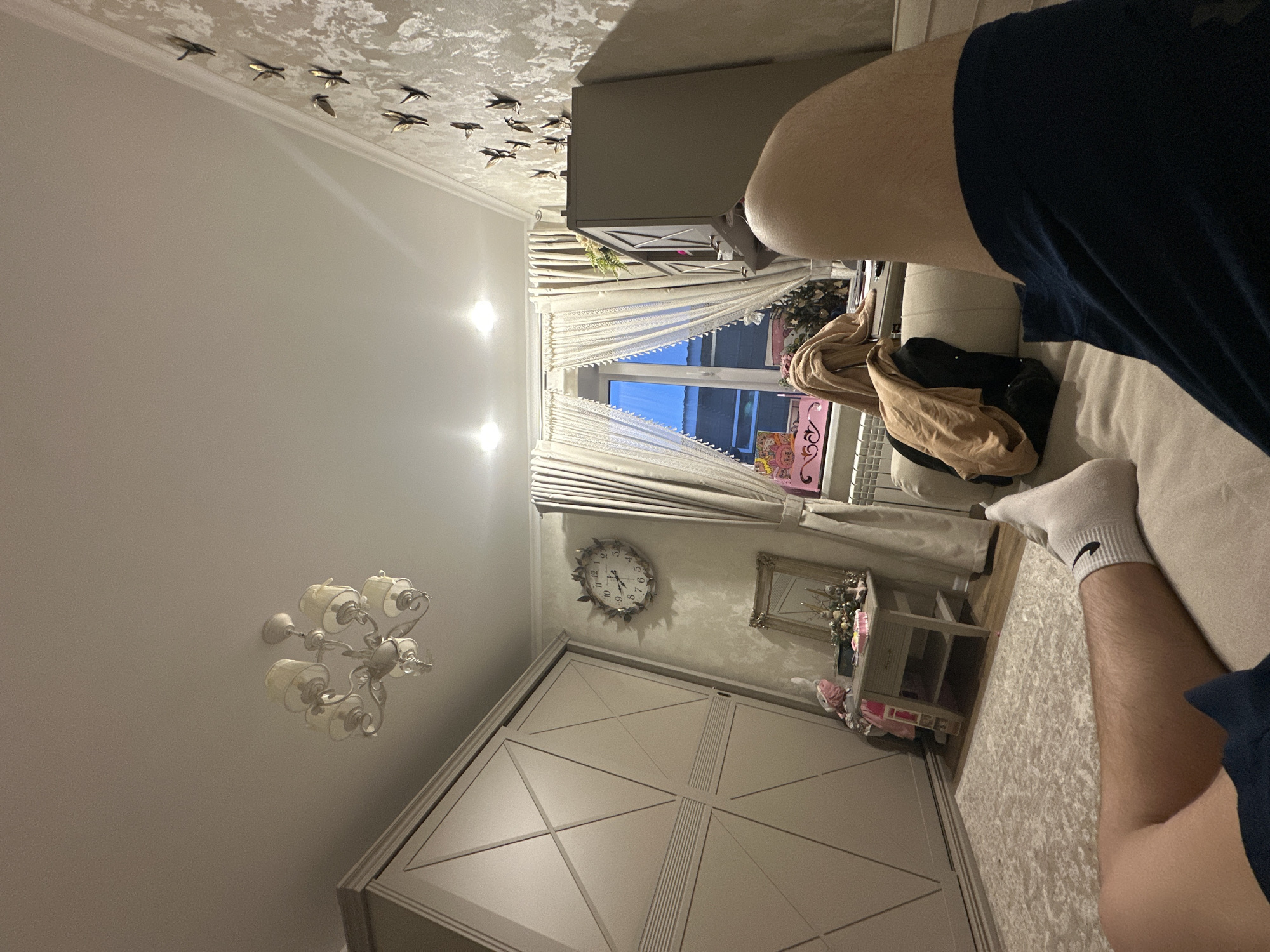 The image shows a view of a room from a low angle. The walls are light-colored with decorative birds affixed to the ceiling. A chandelier hangs from the ceiling. A large wardrobe with a geometric design is visible. A window with white curtains is present. There is a cabinet with decorative items. A clock hangs on the wall. Part of a person sitting on a sofa is visible in the foreground. The person is wearing blue shorts and white socks. The location is an interior space in an unknown city.