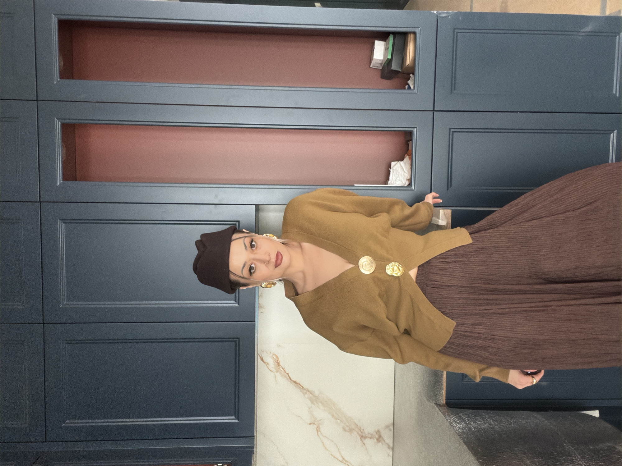 The image displays a young adult woman standing indoors, posing for a photograph. The woman has dark hair, partially covered by a dark brown beret. She wears striking large, gold-toned earrings and dark red lipstick, looking directly at the camera with a confident expression. Her attire consists of a mustard or camel-colored long-sleeved cardigan, which is fastened with two prominent, ornate gold buttons; one features a spiral design, and the other appears to be a relief of a lion's head or a heraldic motif. She is also wearing a dark brown, finely pleated skirt that extends out of the frame.
The background is dominated by floor-to-ceiling cabinets in a deep blue-grey color. Some sections are closed cabinet doors featuring a classic recessed panel design. Other sections are open shelves with a reddish-brown interior, containing various items such as papers, small boxes, and white fabric, suggesting a functional or storage area. Below the woman, a white surface with distinct brown and reddish-brown veining, resembling marble or a similar stone, is visible, likely serving as a countertop or part of the floor. A darker grey surface is visible below this. The lighting is even, typical of an indoor setting. There is no visible text in the image. The location context for this scene is Krasnoyarsk, Russia.