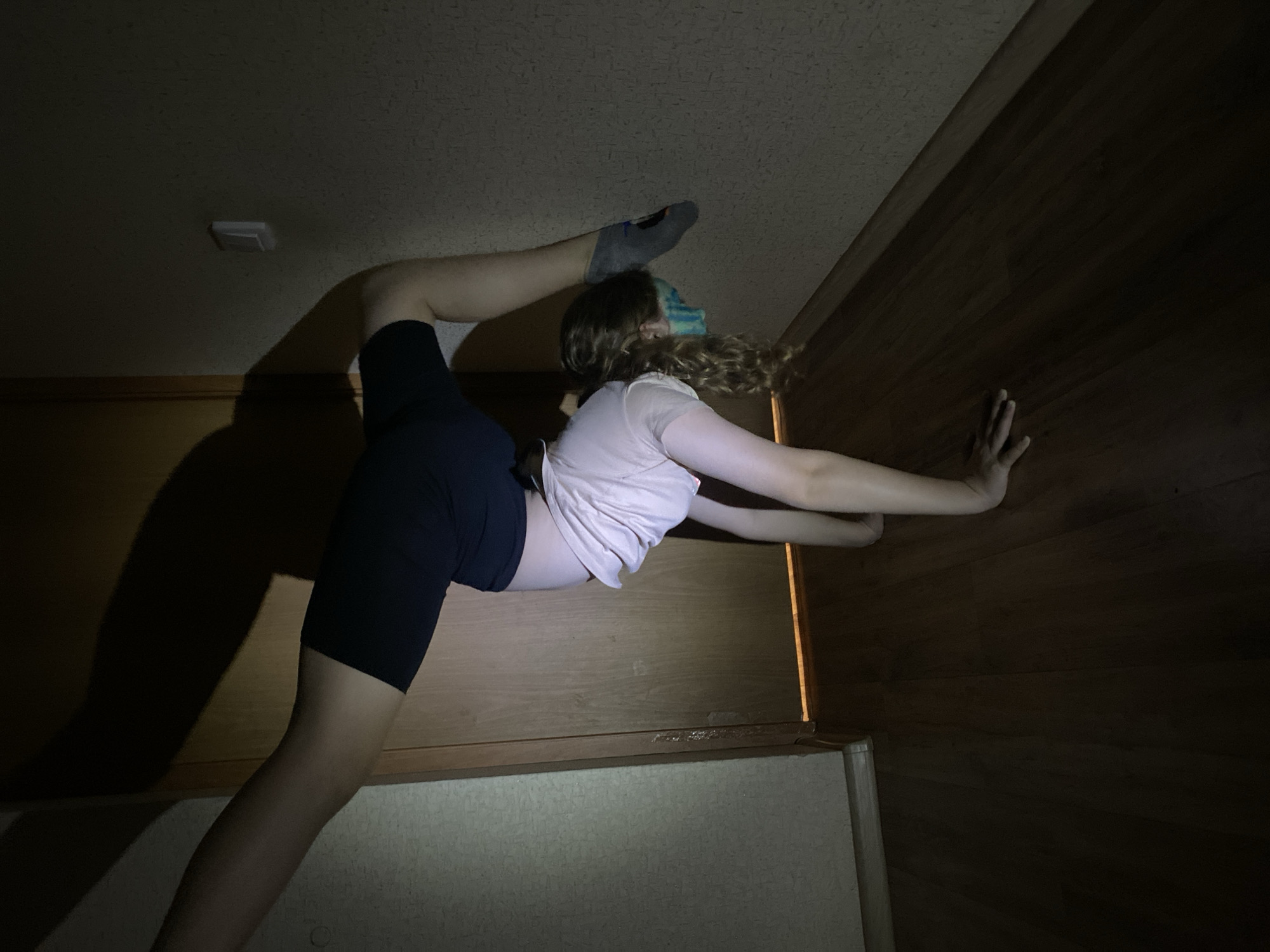 The image shows a person in a dark indoor setting, performing a split against a wall. The person is wearing a light-colored shirt and dark shorts, with one leg extended upwards, the foot touching the ceiling while wearing a sock. The other leg is extended downwards. The person is using one hand to steady themselves against the dark-paneled wall. The room has a ceiling light and a lower wall section in a lighter shade. The location is unknown.