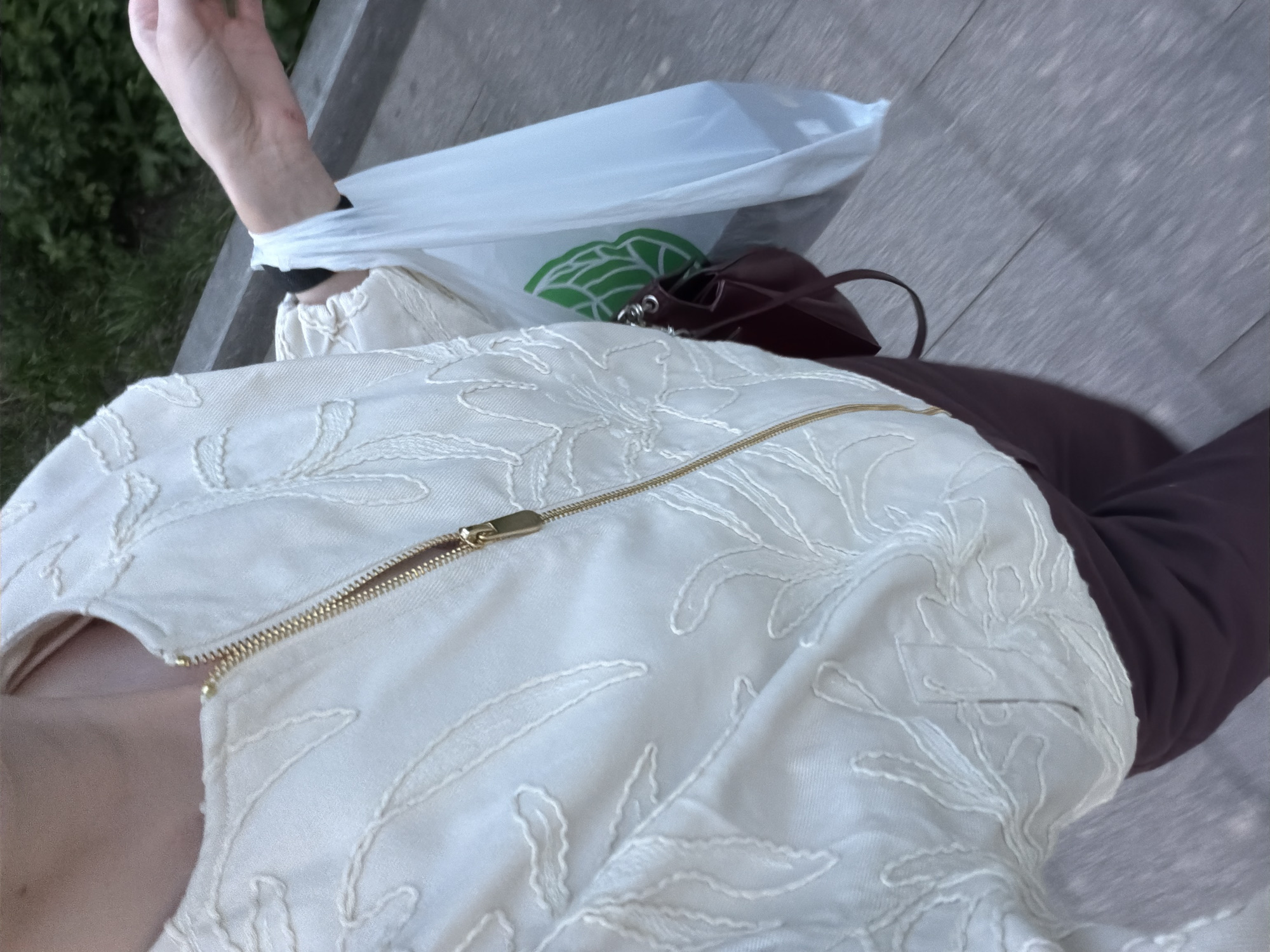 This high-angle image captures a person, likely female, from the chest down, seated outdoors. The individual is wearing a light, possibly cream or off-white, jacket adorned with an intricate, tonal embroidered or embossed leafy pattern. A prominent gold zipper runs vertically along the front of the jacket. Beneath the jacket, a plain dark brown garment, possibly trousers, is visible, along with a hint of a light pink or nude-colored neck/chest area.
One arm is extended upwards and to the left, with a black hair tie or band visible on the wrist. A white plastic shopping bag, featuring a partially visible green stylized leaf logo, is draped over this arm and rests near the person's lap. Adjacent to the plastic bag is a small, dark red or burgundy handbag with silver-toned hardware.
The setting appears to be an outdoor public space, possibly a park or boardwalk, in Moscow, Russia. The person is seated on a light brown wooden surface, consistent with a bench, with blurred green foliage visible in the background on the left. The ground to the right of the person is a light grey, textured surface, resembling pavement or concrete. The lighting suggests daytime, possibly late afternoon or early evening, with soft, diffused illumination. The primary activity depicted is resting or sitting. No legible text is visible in the image.