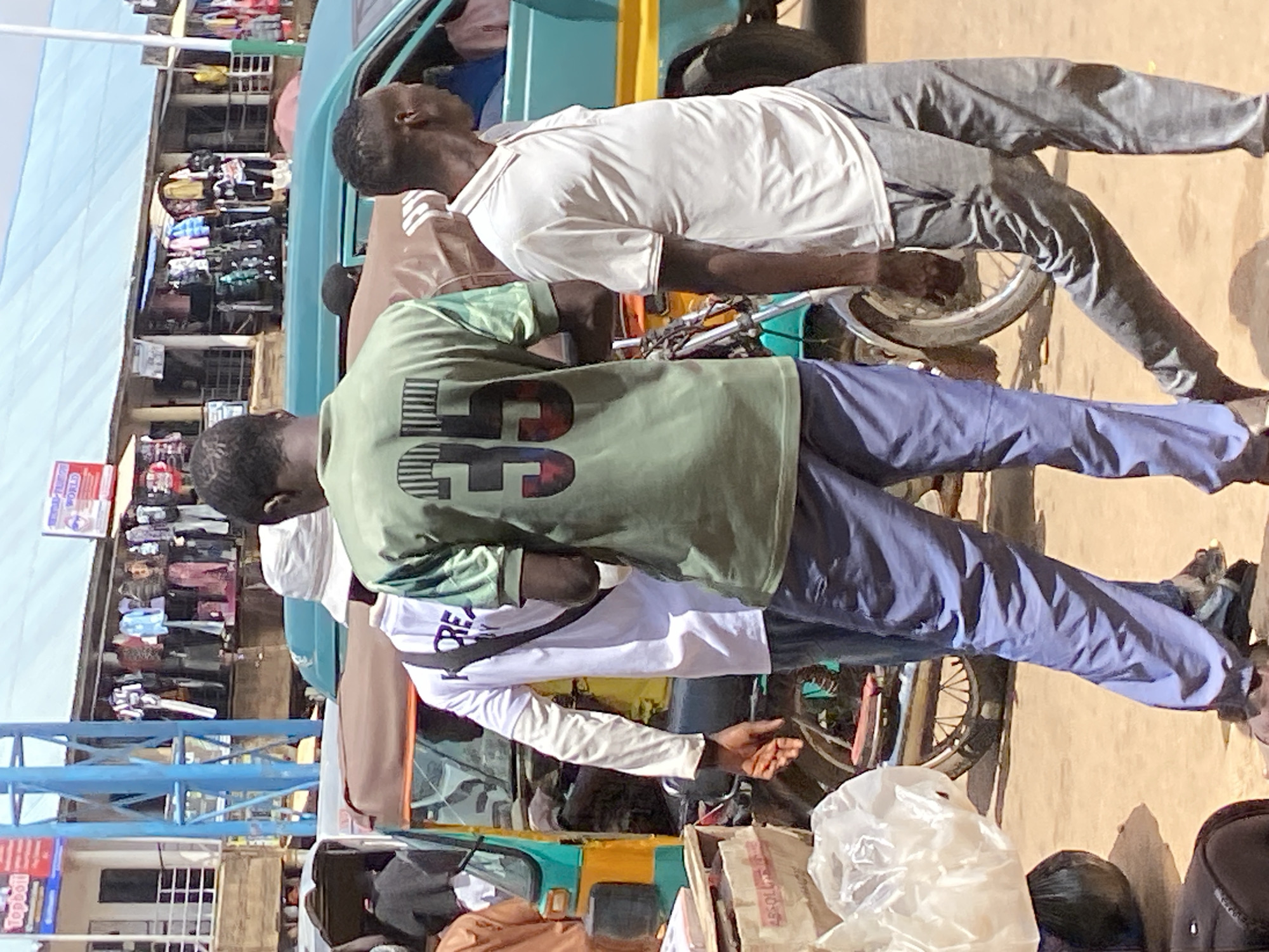 The image depicts a street scene in Bukuru, Nigeria. In the foreground, three men are visible. The man on the left wears a green t-shirt with the number "35" printed on the back in a stylized font. He is also wearing blue pants. Next to him, to the right, another man is partially visible, wearing a white long-sleeved shirt with "KOREA" printed on it, and a black strap across his chest. To the right of the man in the green shirt, a third man is wearing a light-colored polo shirt and gray pants. He is facing to the right.
Behind the men, several vehicles are present. A turquoise and yellow motorized tricycle, commonly known as an "okada" or "kabukabu" in Nigeria, is parked to the left. A teal-colored van or truck is visible behind the men, with a yellow stripe along its side.
The background reveals a marketplace. Stalls with clothing displayed for sale are lined up under a blue corrugated metal roof. Various items of clothing, including shirts, pants, and dresses, hang on racks. Signs with text are visible above the stalls, suggesting commercial activity. The ground appears to be unpaved, dusty earth. The overall impression is that of a busy, outdoor market environment.