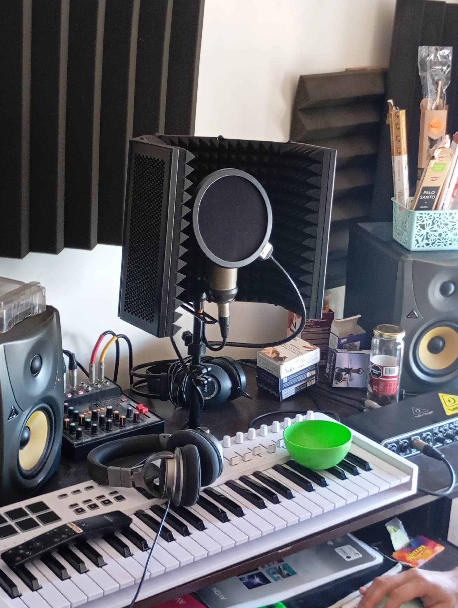 This media file captures a close-up, eye-level view of a home studio or creative workspace, primarily set up for audio production or recording. The scene is illuminated by natural light, suggesting it is daytime.
The central subject is a condenser microphone, equipped with a circular pop filter, enclosed within a portable sound shield or vocal booth, indicating an emphasis on vocal recording. The microphone is mounted on a stand. To the left, a studio monitor speaker with a prominent yellow cone is visible, connected to a small audio mixer via red and yellow cables. Another similar speaker is partially visible on the right. A pair of over-ear headphones rests on a white MIDI controller keyboard, which features black and white keys. A black remote control with a partially visible Netflix logo is also on the keyboard.
Cluttering the desk are several miscellaneous items: a light blue, patterned container holding sticks of incense, with "PALO SANTO" and "HEM" clearly legible on the packaging. Nearby, small boxes, some labeled "Meditation," are stacked. A glass jar with a gold lid, containing an unidentifiable substance, displays a barcode and Spanish text including "Peso Neto". A vibrant green bowl sits on the MIDI keyboard. Under the right speaker, a Behringer audio interface is partially visible. Beneath the desk, a book or folder with the "AVON" logo can be seen.
In the bottom right corner, a person's hand is visible, resting on a device that appears to be a laptop or a trackpad, suggesting active engagement with the studio setup. Acoustic foam panels are visible on the wall behind the equipment, indicating an attempt to manage sound reflections. The presence of Spanish text on packaging and the type of items are consistent with the given context of Buenos Aires, Argentina. The overall impression is a functional, personal recording or music production space.