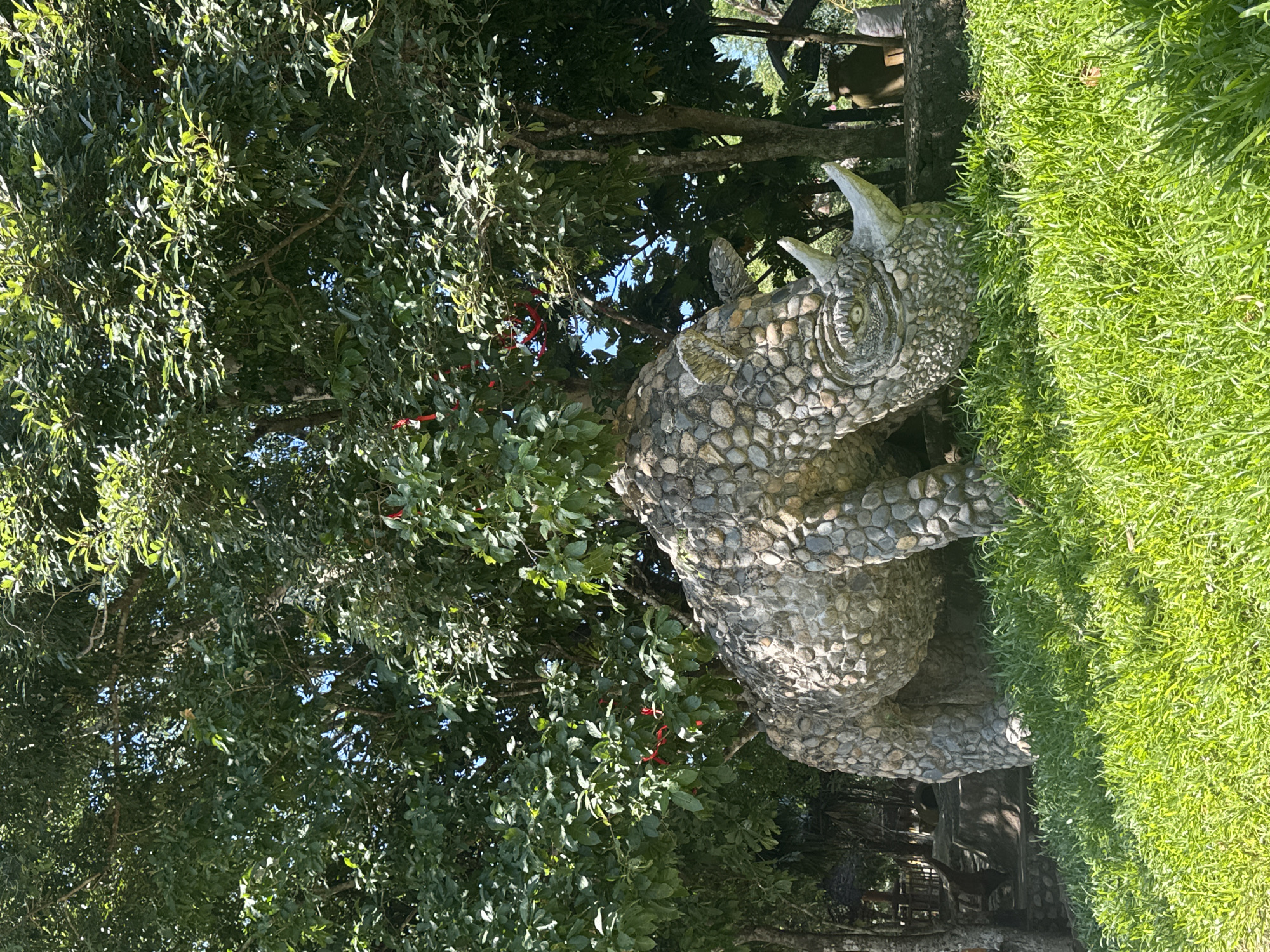 A statue of a rhinoceros, constructed from rough, gray stones, is positioned in a grassy area under the shade of lush green trees. The rhinoceros is facing downwards, as if grazing. Its details, including its horn, eyes, and skin texture, are clearly visible, all rendered in stone. In the background, more trees and some hints of structures suggest an outdoor location, possibly a park or a zoo. The lighting indicates it is daytime, with sunlight filtering through the leaves. There are no people present in the scene. The location is identified as Nam Khanh Vinh, Vietnam.