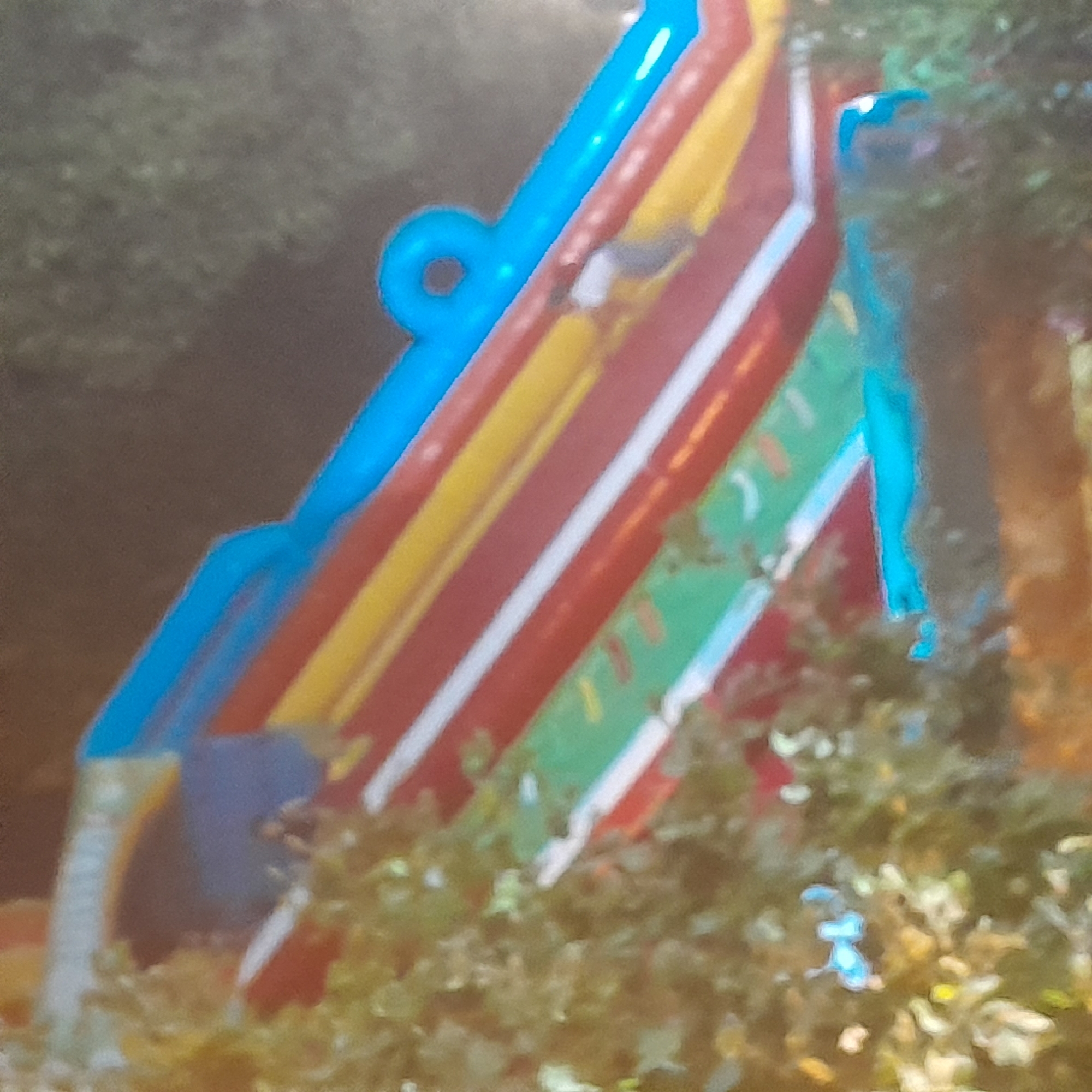 The image shows a close-up, blurred view of a brightly colored inflatable structure, possibly a water slide or bouncy castle, against a dark, out-of-focus background that suggests an outdoor setting at night or in low light. The inflatable features prominent horizontal stripes of blue, yellow, red, and green, with white trim separating some of the colors. A person, appears to be in the upper right portion of the slide, wearing dark clothing. The overall scene suggests a recreational or entertainment venue. The location in Rudəkənar, Azerbaijan is not visually discernible from the provided image.