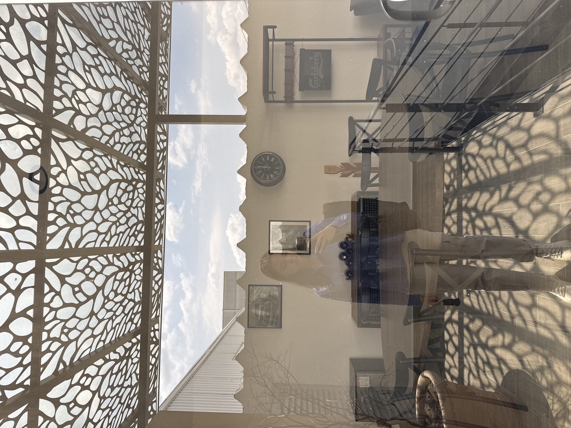 The image, possibly taken from inside a building in San Bernardino Tlaxcalancingo, Mexico, shows a complex scene with multiple layers. On the left is an ornate, patterned metal screen or wall. Through the window, the sky with clouds is visible, and part of a building with a ridged roof. Reflections on the glass create a layered effect, revealing the interior of the building: a person is visible, as well as a shelf with wine bottles, wall decor including pictures and a clock, furniture, and a Carlsberg sign. The shadows of the patterned screen are cast on the floor.