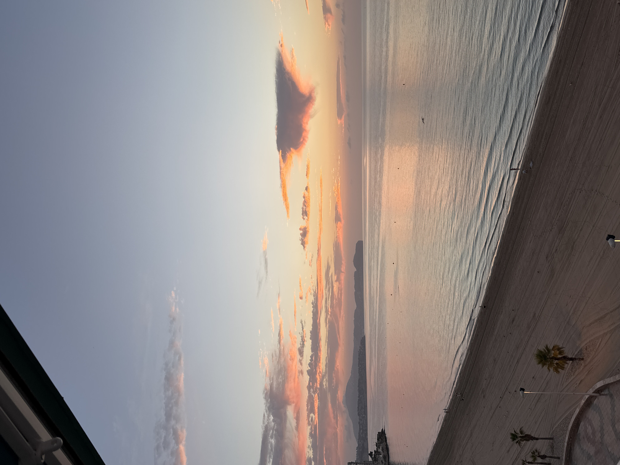 The image depicts a serene sunrise over the Mediterranean Sea in Calp, Spain. The sky is a gradient of soft blues and pinks, with scattered clouds catching the early morning light. The calm sea reflects the colors of the sky, creating a tranquil atmosphere.
In the foreground, a sandy beach stretches along the coastline. Several palm trees are scattered across the beach, and the sand is marked with footprints and tire tracks, suggesting recent activity. A few people can be seen walking along the shore, and a lone bird soars in the distance. A curved, paved walkway lines the edge of the beach.
In the background, the Calpe coastline is visible, with a prominent rock formation rising from the sea. Buildings are clustered along the hills, and the landscape is bathed in the soft glow of the sunrise. The overall scene is one of peace and natural beauty.
