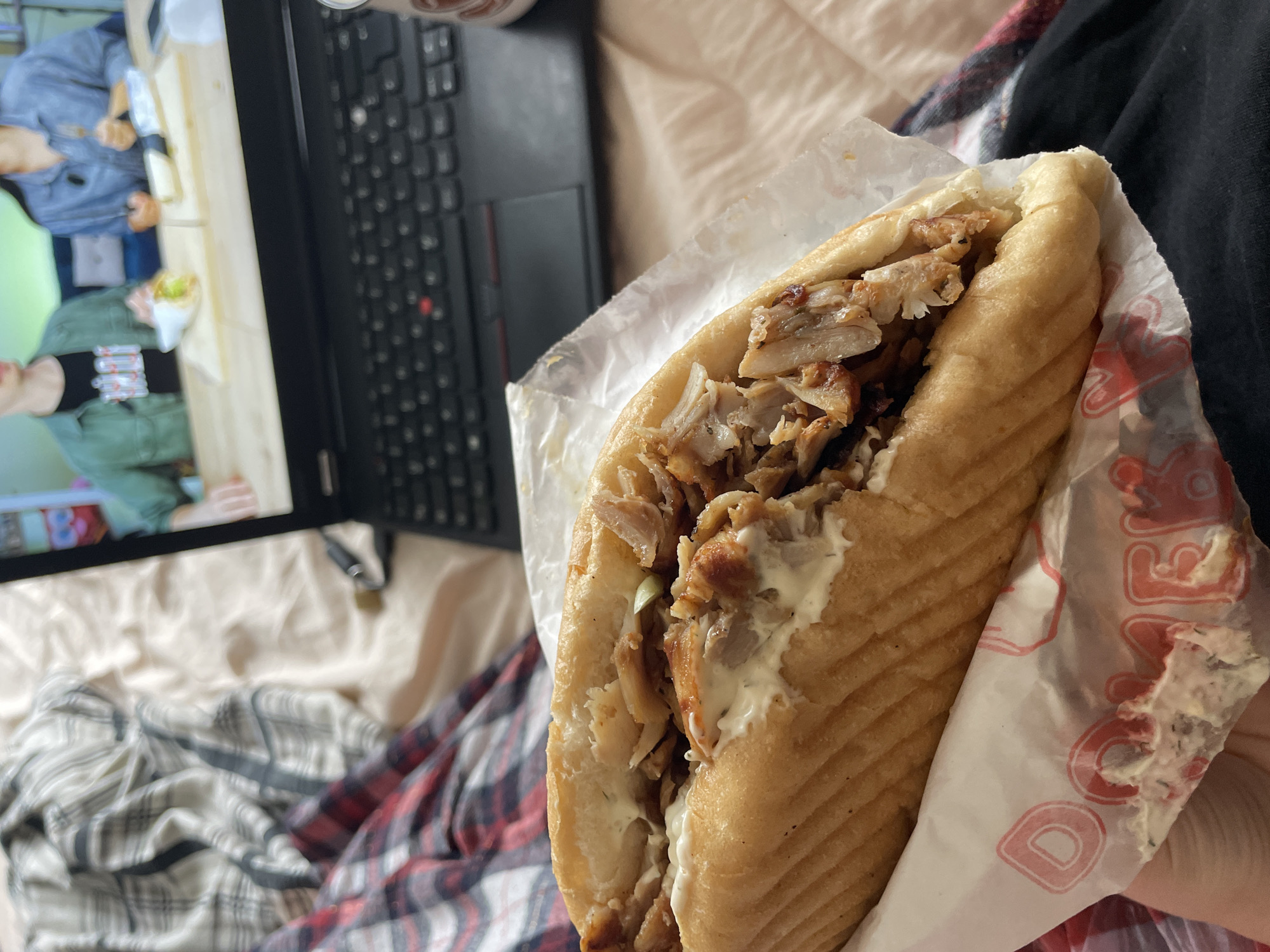 A close-up shot shows a partially eaten döner kebab in a paper wrapper, held in someone's hand. The filling includes meat and a white sauce. In the background, out of focus, is a laptop displaying a video. The surrounding environment includes bedding and a plaid blanket. There is no evidence to indicate the location is Zlatograd, Bulgaria.