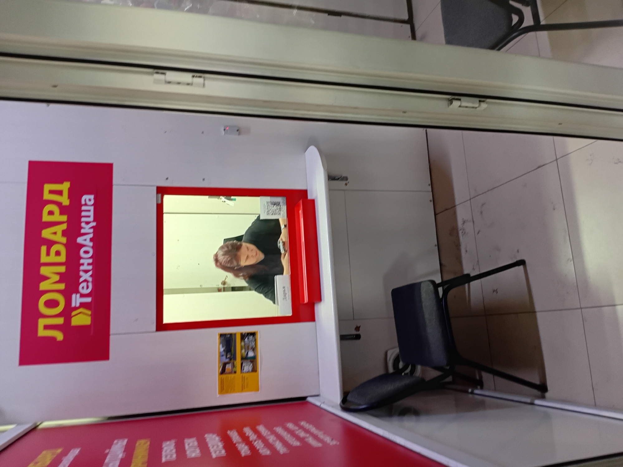 A young woman with dark hair pulled back is seated behind a counter in what appears to be a pawn shop or loan office in Semey, Kazakhstan. She is looking down at a mobile device in her hands, which she is holding on the counter. The counter is white and curves slightly. Above the counter, a windowed opening displays a bright, light-colored interior. A red frame surrounds the opening, and a red tray sits on the counter below the opening. A sign above the window reads "ЛОМБАРД ТехноАкша" in yellow and white letters against a red background. To the left of the counter, a black chair with a metal frame is positioned. A red wall with white text is visible on the far left of the image, partially obscured. The floor is tiled in a light color. The overall scene suggests a typical commercial establishment.