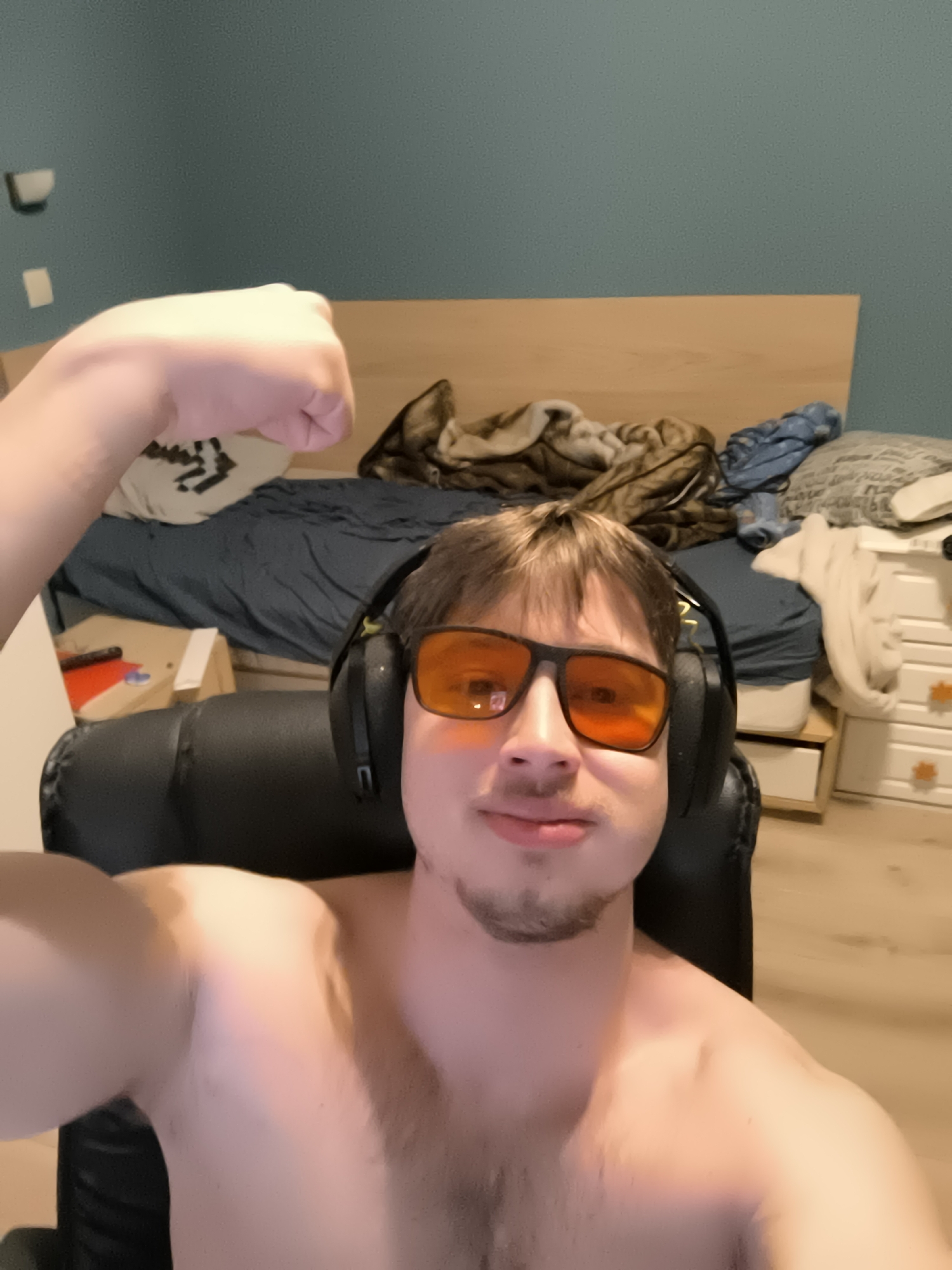 A shirtless, fair-skinned person with short brown hair is seated in a black chair, wearing black headphones and orange-tinted glasses. They are flexing their right arm in a selfie. The background shows a bed with a dark blue sheet, a wooden headboard, pillows, and rumpled blankets. To the right of the bed is a white dresser with orange knobs. The wall behind the bed is blue. A light switch is visible on the upper left wall. The flooring is light-colored wood. There is no evidence to suggest this photo was taken in Sofia, Bulgaria.