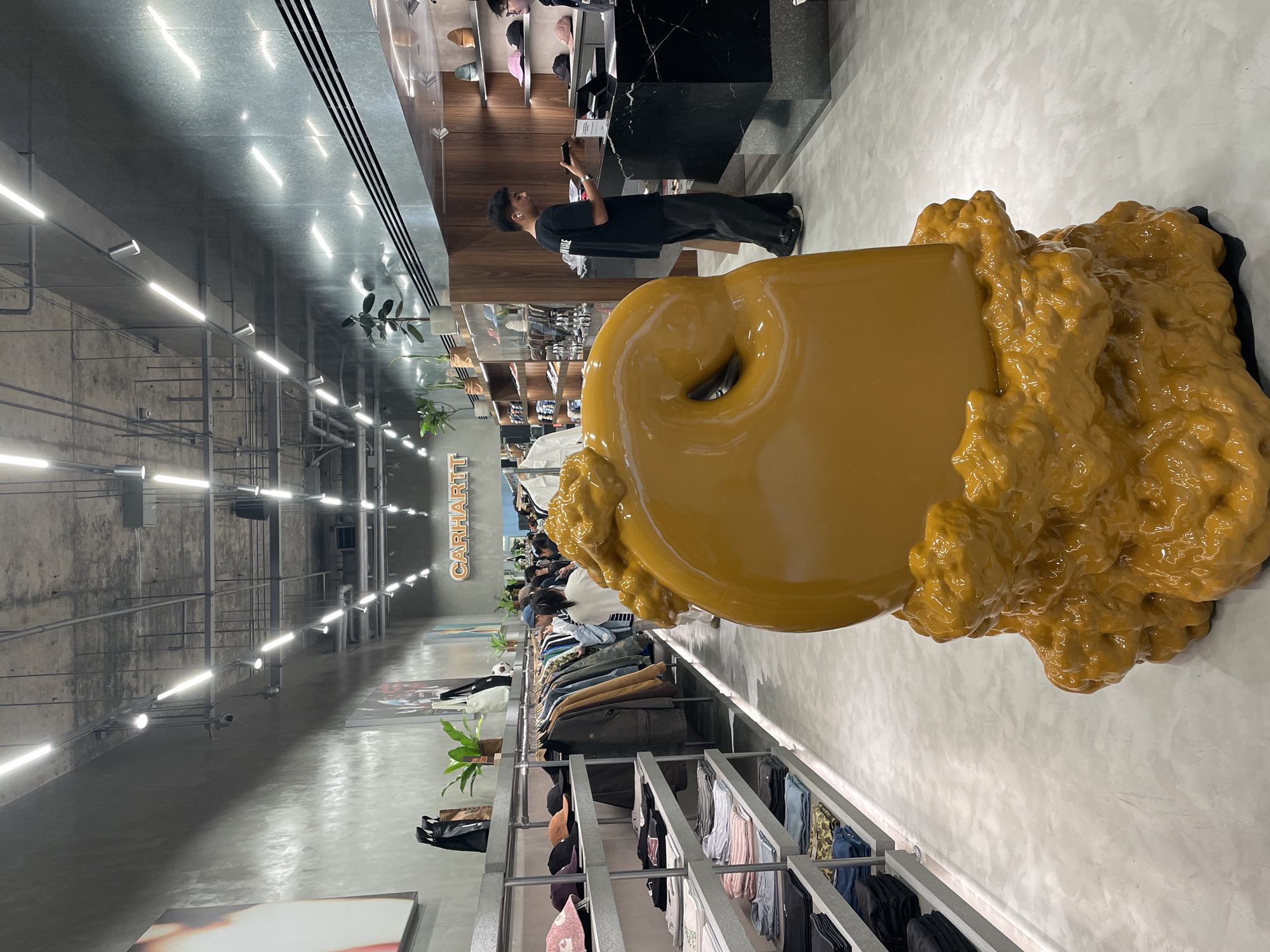 The image captures the interior of a modern retail store with an industrial-chic aesthetic. In the right foreground, a large, glossy mustard-yellow sculpture dominates the space. This abstract artwork features a smooth, rounded main body with a central, ring-like indentation, and an irregular, textured mass that appears to flow or melt, suggesting an organic or dynamic form.
Further into the store, slightly to the left of the sculpture, a person dressed in a black t-shirt and dark pants stands at a sleek black counter with a marble top, engrossed in their smartphone. Behind this counter, shelves display a variety of caps and hats. The store features multiple clothing racks filled with diverse apparel, including jackets, shirts, and pants, which are being browsed by several other shoppers visible in the background.
The architecture of the store includes concrete walls with visible piping and conduits, contributing to an urban, industrial feel. The ceiling is reflective, adorned with long, parallel lines of fluorescent tube lights and individual spotlights that brightly illuminate the entire area. Green potted plants are interspersed throughout the store, adding natural elements to the contemporary design. On a wall in the background, the lighted text "CARHARTT" is visible, identifying the brand. The scene depicts a well-lit, active retail environment during what appears to be daytime. The location context is Bangkok, Thailand.