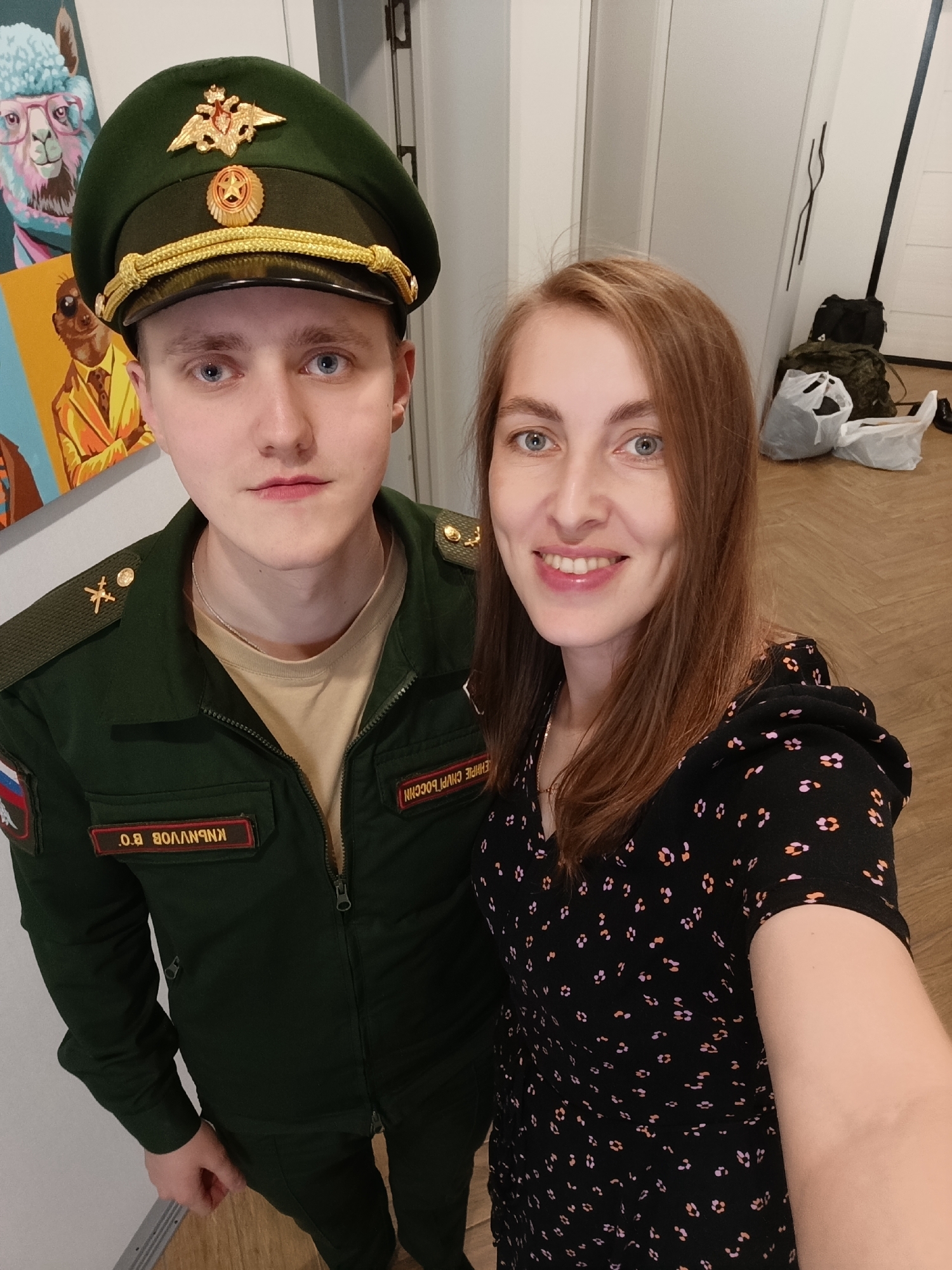This media file shows a close-up, selfie-style shot of a young man and a woman. The man is on the left, wearing a dark green military uniform with a matching peaked cap. The cap has gold insignia, including an emblem with wings and a star, and a gold cord band. The uniform jacket also has gold insignia on the shoulders and a patch with the Russian flag and lettering on the left chest. A red name tag on the jacket reads "О.В. ВОЛЧАНКИН" in white Cyrillic letters, and another patch on the right chest reads "Е.М.Р.Е. РОССИИ". He has fair skin, light blue eyes, and light brown hair.
The woman is on the right, with light brown hair that falls over her shoulders. She is smiling and looking towards the camera. She is wearing a black dress with a small, colorful floral pattern. Her skin is fair, and she has blue eyes.
The background appears to be indoors, possibly in a hallway or room with wooden flooring. To the right, there are white doors and some bags, including a green backpack and plastic bags. The lighting suggests it could be daytime. The overall mood appears positive and casual, as they are posing for a picture together. The visible text on the man's uniform indicates he is affiliated with the Russian military. The location context provided is Chaikovsky, Russia.