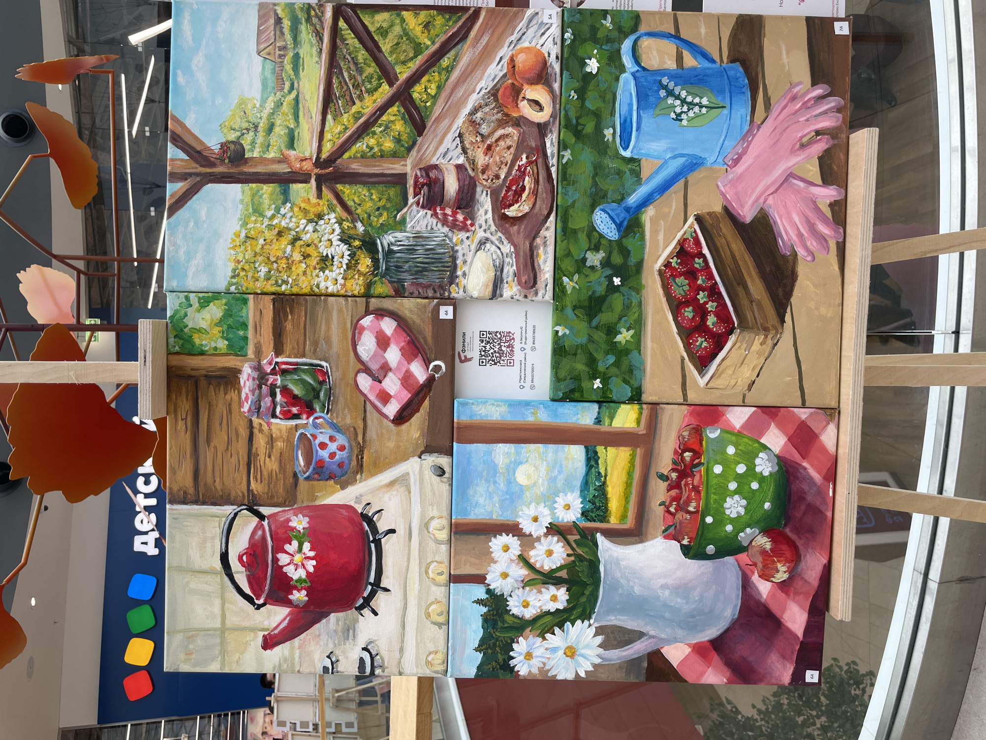 The image displays a collection of six distinct paintings, likely part of an art exhibition, mounted on a wooden easel. The paintings are arranged in a 2x3 grid. The background shows a blue wall with the word "ДЕТСКИЙ" (Children's) in large white letters, suggesting a children's store or cultural center. There are also some abstract orange decorations hanging from the ceiling and metal shelving units in the background. The bottom right portion of the image reveals a partial view of a glass storefront with reflections of the surroundings, including an indication of a city location.
The paintings themselves depict various still life and landscape scenes:
* **Top Left:** A red teapot with white floral decorations sits on a stovetop. To its right, a blue mug with red polka dots and a jar of pickled cucumbers are visible. A red and white checkered oven mitt lies nearby.
* **Top Right:** A rustic outdoor scene with a wooden railing overlooking a verdant landscape with a farmhouse in the distance. A ginger cat is perched on the railing, and a basket of yellow flowers hangs from the porch.
* **Middle Left:** A white pitcher filled with white daisies stands on a red and white checkered tablecloth. A green bowl overflowing with red apples and a single apple on the side are also depicted.
* **Middle Right:** A wooden cutting board displays a loaf of bread, a jar of jam, a pat of butter, and sliced fruit (likely peaches or apricots). A white napkin or tablecloth is spread beneath the items.
* **Bottom Left:** A wooden crate filled with bright red strawberries is placed on a wooden surface.
* **Bottom Right:** A blue watering can with a Lily of the Valley motif is next to a pair of pink gardening gloves.
The overall impression is a vibrant display of artwork, likely at a public venue in the city of Perm, Russia, given the signage. The lighting suggests an indoor setting with ambient artificial light. No people are clearly visible interacting with the artwork, though blurred figures are faintly discernible in the background through the glass.