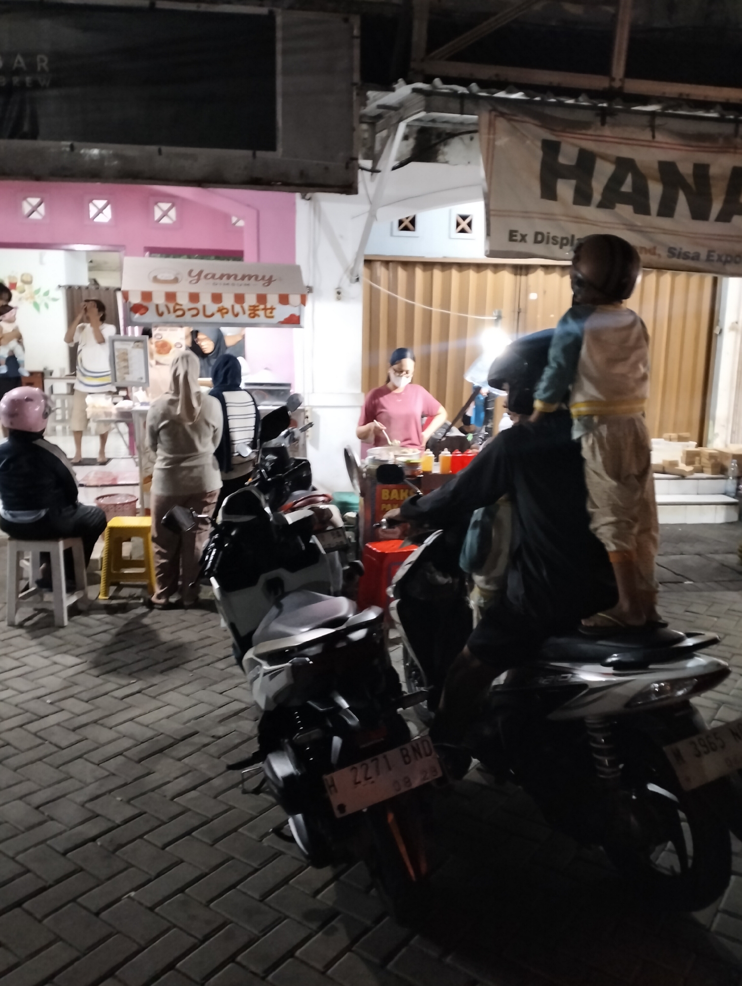 The image depicts a nighttime street scene in Semarang, Indonesia, likely in an area with food vendors. In the foreground, two motorcycles are parked on a patterned brick pavement. One motorcycle, in the middle of the frame, has two people on it. The person in the front, wearing a black shirt and helmet, is seated. The person in the back is standing on the motorcycle's seat, wearing a light-colored jacket and trousers, and a helmet. The second motorcycle, to the left, has a person wearing a black jacket and a pink helmet seated on it.
Behind the motorcycles, several food stalls are visible. One stall has a sign with Japanese text, "いらっしゃいませ" (Irasshaimase), meaning "Welcome," and a brand name "Yammy." Another stall has people working behind it, preparing food. A banner with the word "HANA" is hanging above the stalls. The background shows building facades and lighting. The overall atmosphere suggests a busy, outdoor market or food court.