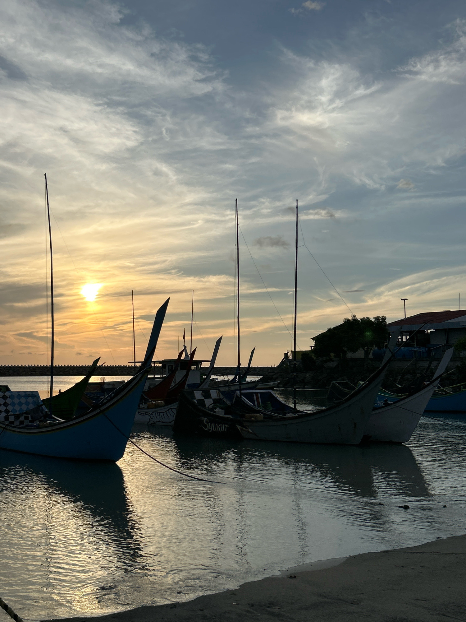 The image depicts a harbor scene in Sabang, Indonesia, at sunset. Several traditional fishing boats with distinctive pointed prows are moored in calm water, their reflections shimmering on the surface. The sky is filled with dramatic clouds, illuminated by the setting sun which is visible as a bright orb on the left side of the frame. The boats are silhouetted against the warm glow of the sunset and the cooler tones of the sky. The shoreline in the foreground consists of sand and a dark line of what appears to be a concrete barrier or road. In the background, a few buildings and trees are dimly visible on the land. The overall impression is one of tranquility and the end of a day's work for the fishermen.