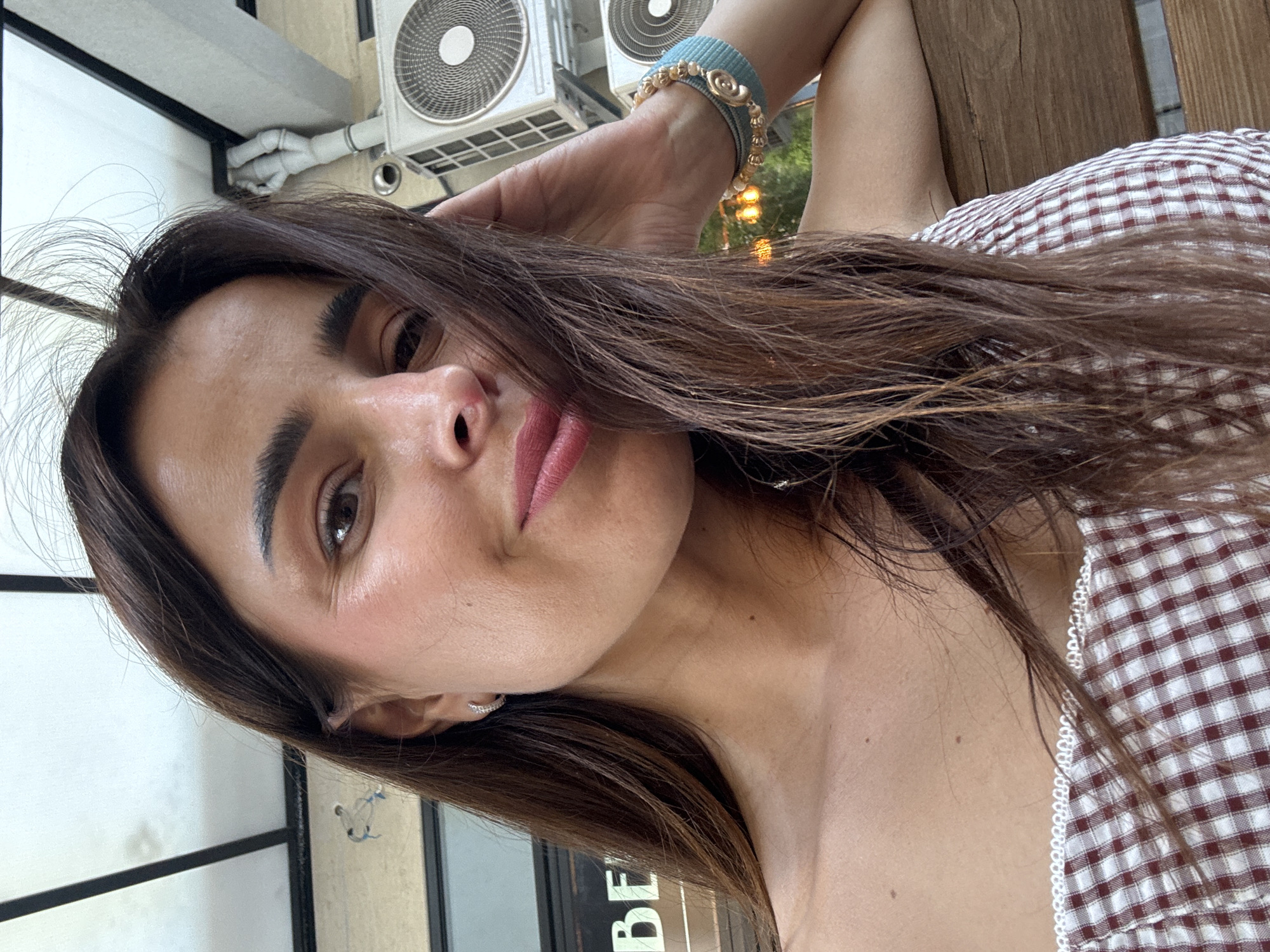 A woman with long, dark brown hair is captured in a close-up, outdoor, selfie-style shot. She is wearing a red and white checkered off-the-shoulder top and a light blue bracelet with gold accents on her left wrist. Her right arm is bent, with her hand resting on her head and near her hair. She has makeup on, including pink lipstick and blush. In the background, there are two air conditioning units mounted on a wall, a wooden bench or table, and what appears to be some greenery and lights, suggesting an outdoor seating area of a cafe or restaurant in Podgorica, Montenegro. The overall lighting is bright and natural.