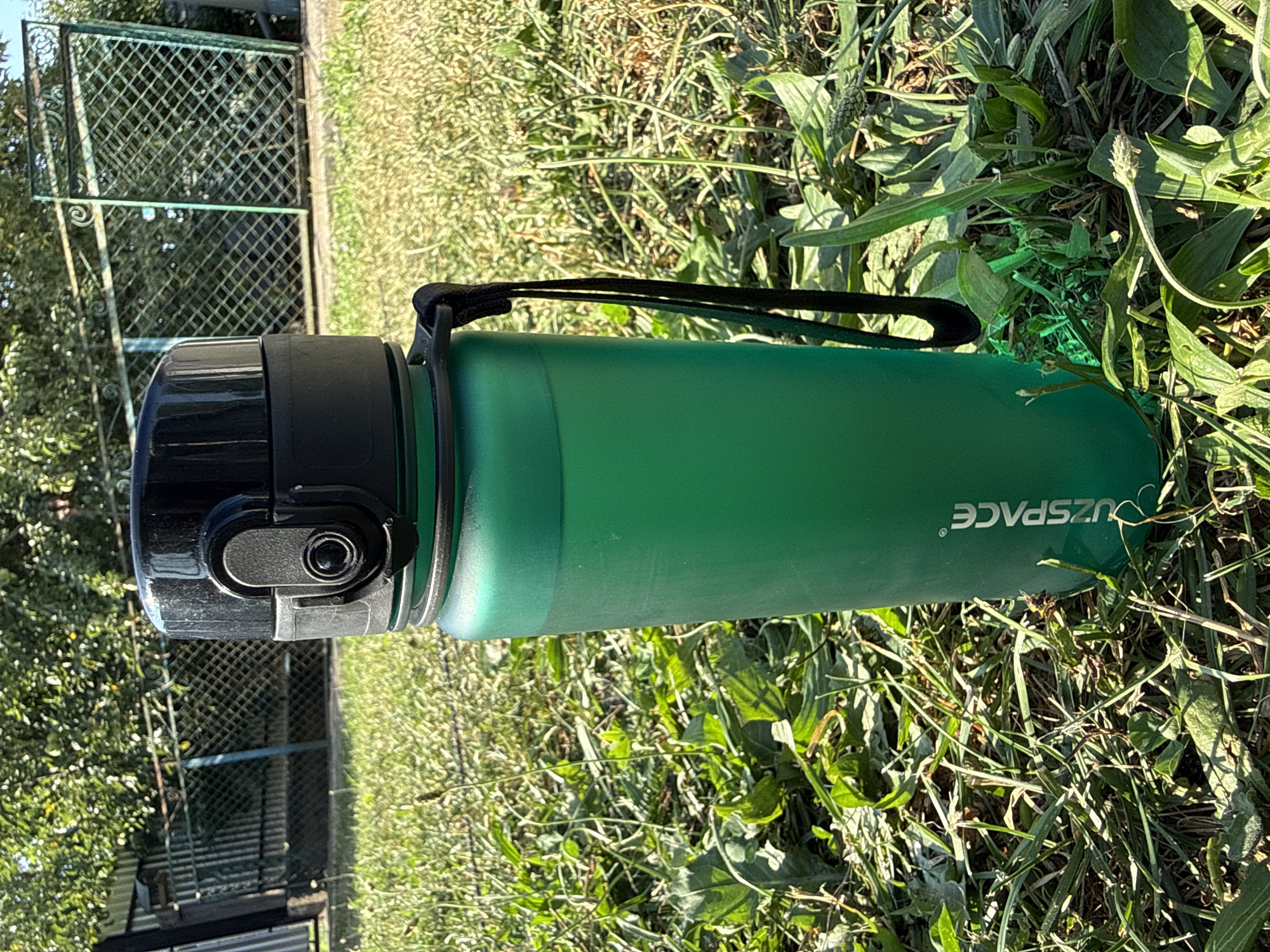 A tall, matte green water bottle with a black lid stands upright on a grassy surface. The bottle has a black strap attached to its lid. The lid features a gray flip-up spout with a black button. The bottle's base is obscured by grass. The background shows a green, ornate metal gate and a fence, with trees and foliage behind them. The scene is illuminated by bright sunlight, casting shadows. The words "UZSPACE" are printed vertically in white on the side of the bottle.