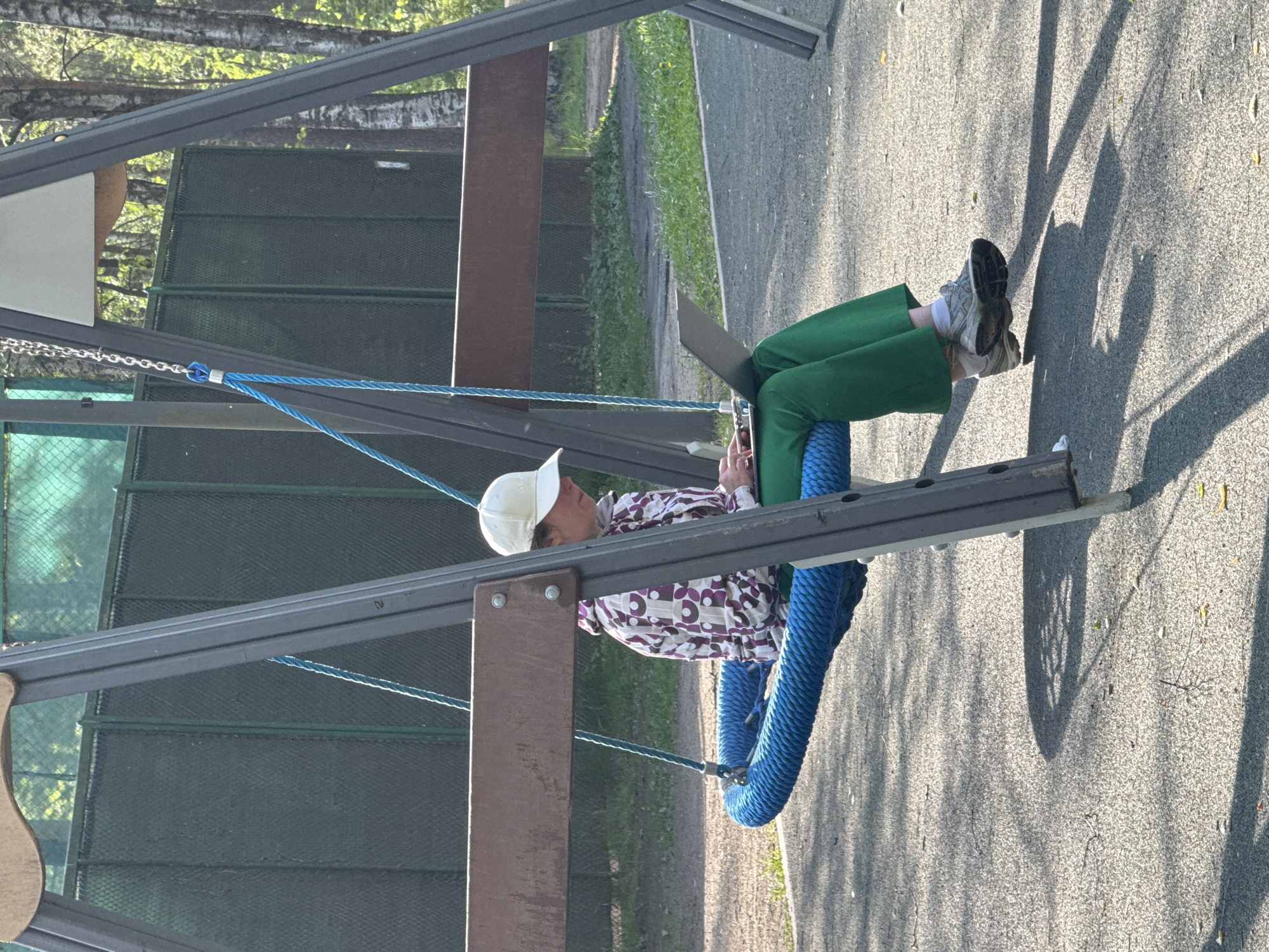 A person sits on a blue tire swing in an outdoor playground in an unknown city in Russia. The individual wears a white baseball cap, a printed long-sleeved shirt, and green pants, and appears to be using a smartphone. The playground has a green metal mesh fence, asphalt flooring, and metal swing set supports. Light filtering through the trees casts shadows on the ground.
