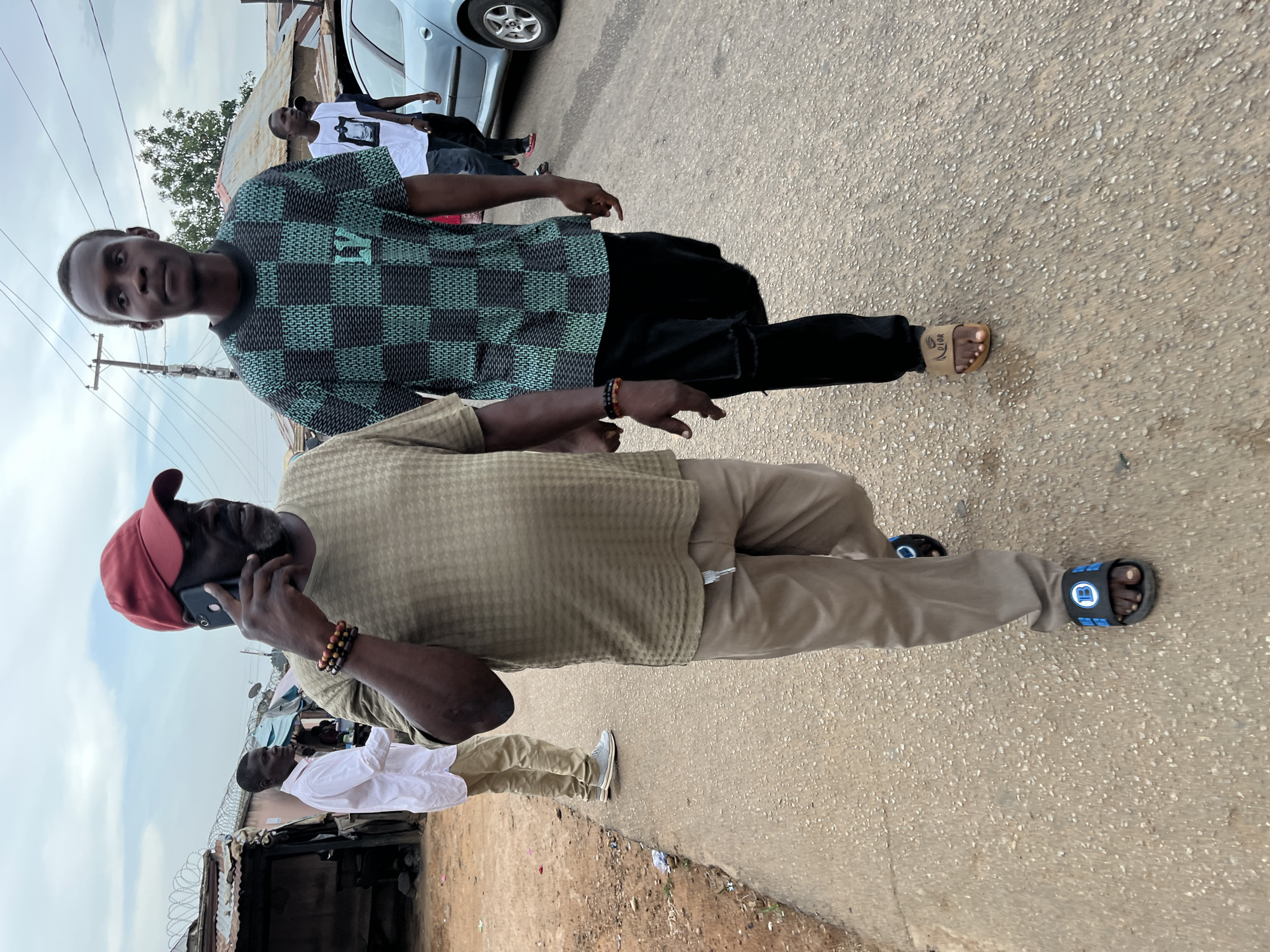 The image shows two men walking along a paved road in Jos, Nigeria. The man closer to the camera is wearing a tan waffle-weave shirt, khaki pants, a red baseball cap, and black sandals with blue accents. He is holding a black smartphone to his ear, engaged in a phone call. The other man is wearing a green and black checkered shirt, black pants, and light brown Dior sandals. There are other people visible in the background, along with a silver car. The environment consists of a city street with a dirt shoulder on one side, and buildings or structures visible in the background, including a building with a corrugated metal roof with barbed wire on top. Power lines are also visible overhead.