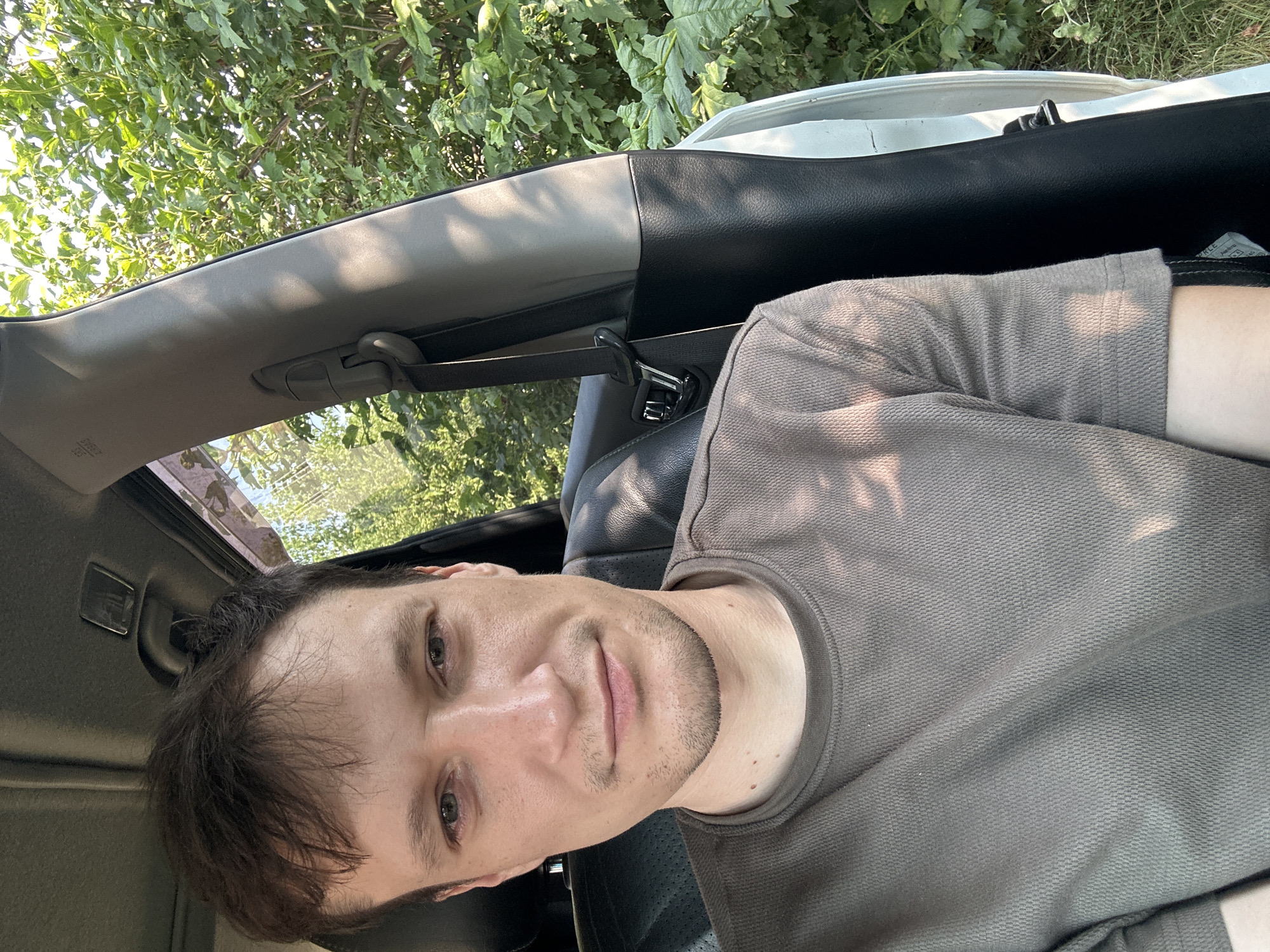The image shows a close-up, waist-up selfie of a man taken inside a vehicle. He is seated in the driver's seat, facing the camera with a neutral expression. He has short, dark brown hair and light-colored eyes. He is wearing a textured, olive-green t-shirt. The interior of the car is visible, including the seat and a portion of the door panel and pillar. Through the open car door and windows, lush green foliage and trees are seen, suggesting an outdoor, possibly rural, setting. There is no discernible indication of the specific location within Shatava, Ukraine.